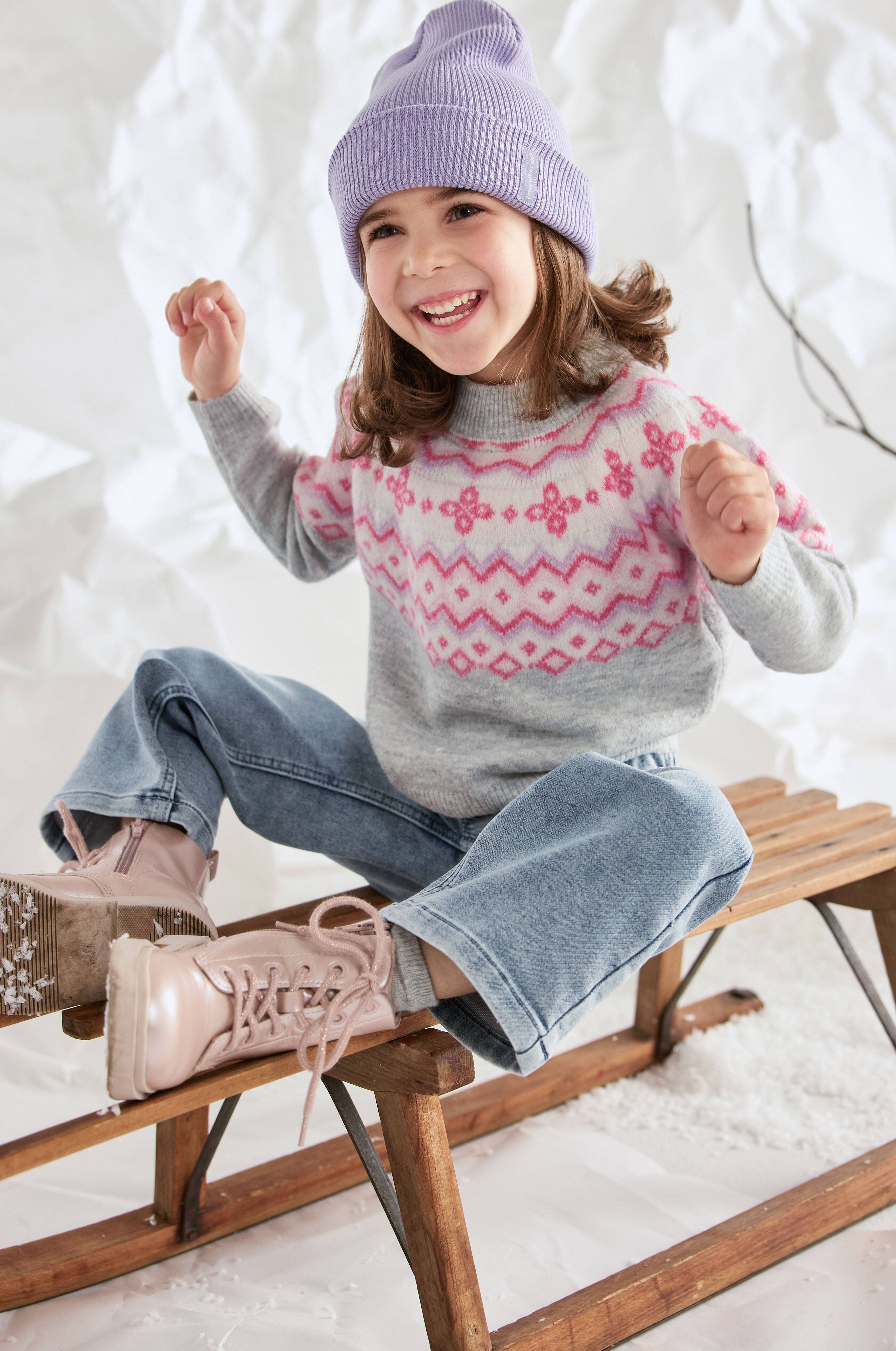 Fille avec un bonnet violet, un pull tricoté à motif nordique et un jean large, assise sur une luge en bois.