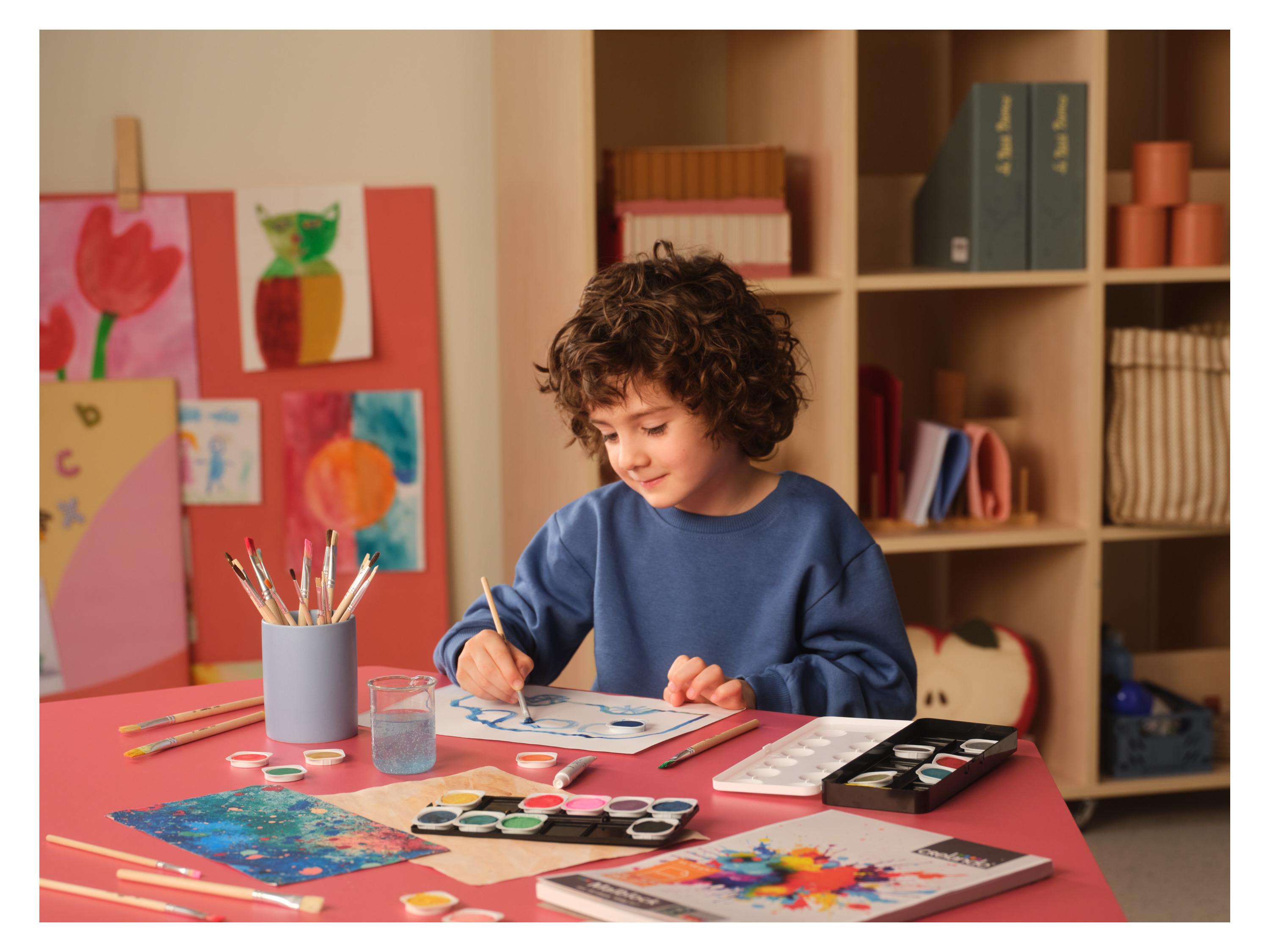 Un enfant peint avec des pinceaux et des aquarelles sur une table rouge, entouré de dessins.
