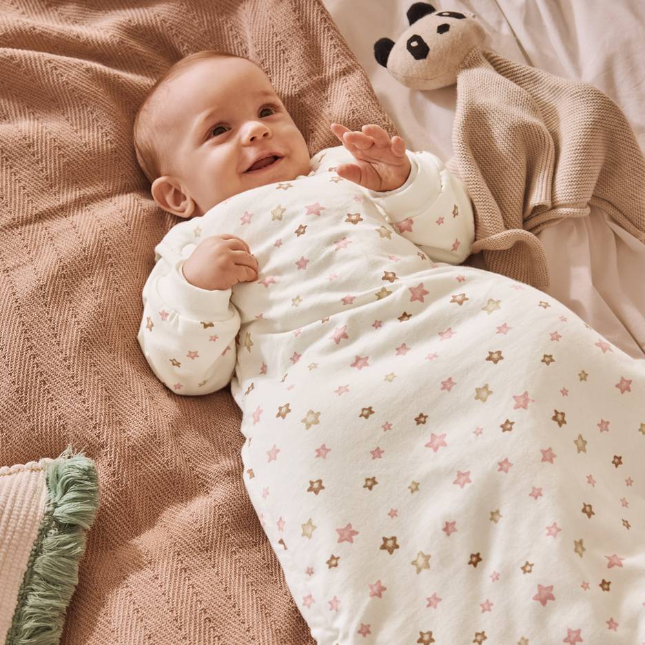 Bébé souriant dans une gigoteuse blanche à étoiles roses et marron, allongé sur une couverture.