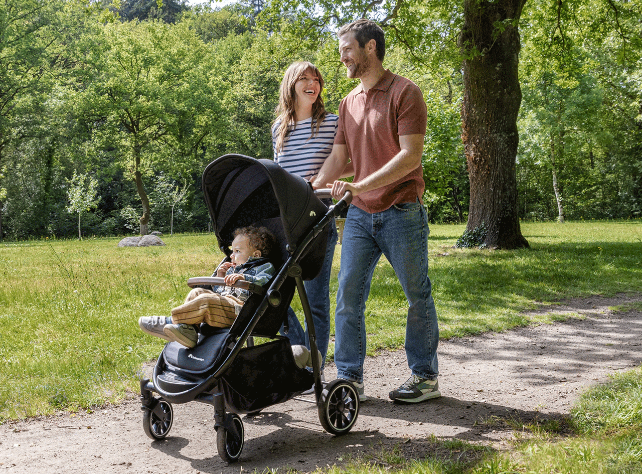 Poussette avec un bébé, poussée par un couple souriant dans un parc verdoyant.