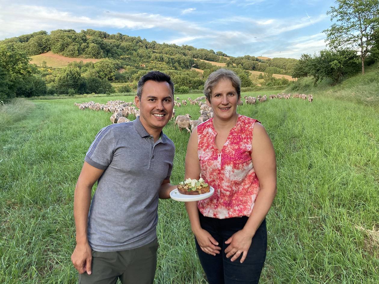 Deux personnes souriantes dans un champ avec un troupeau de moutons, l'une tenant une assiette de nourriture.