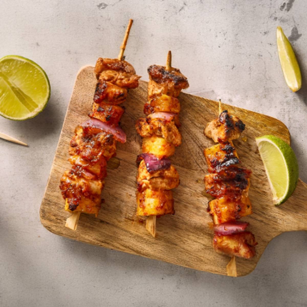 Trois brochettes de poulet grillé marinées, servies avec des quartiers de lime sur une planche à découper en bois.