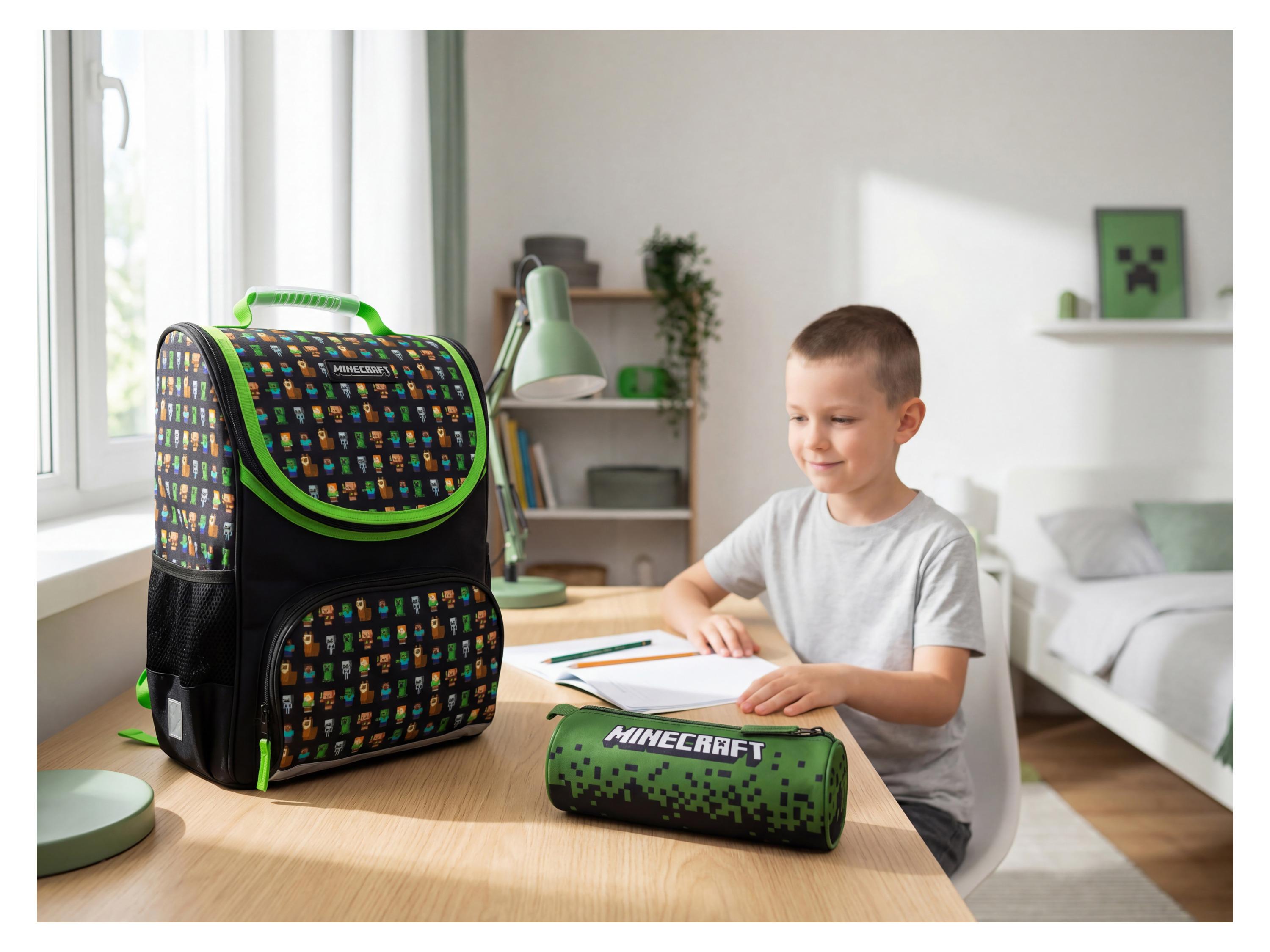Un enfant souriant dessine à un bureau avec un sac à dos et une trousse à motifs de personnages de jeu vidéo MINECRAFT
