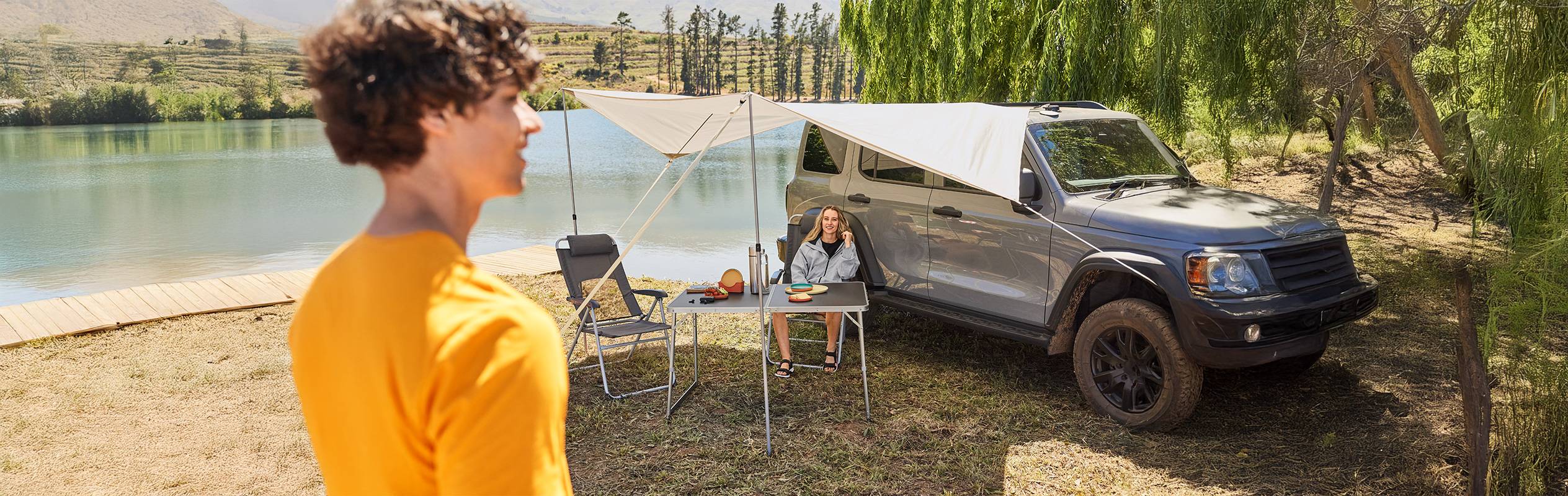 Un homme et une femme campent au bord d'un lac avec un auvent de voiture et une table de camping.