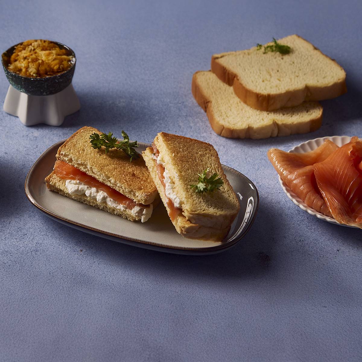Sandwichs au saumon fumé et fromage frais, avec du pain de mie et un bol de garniture.