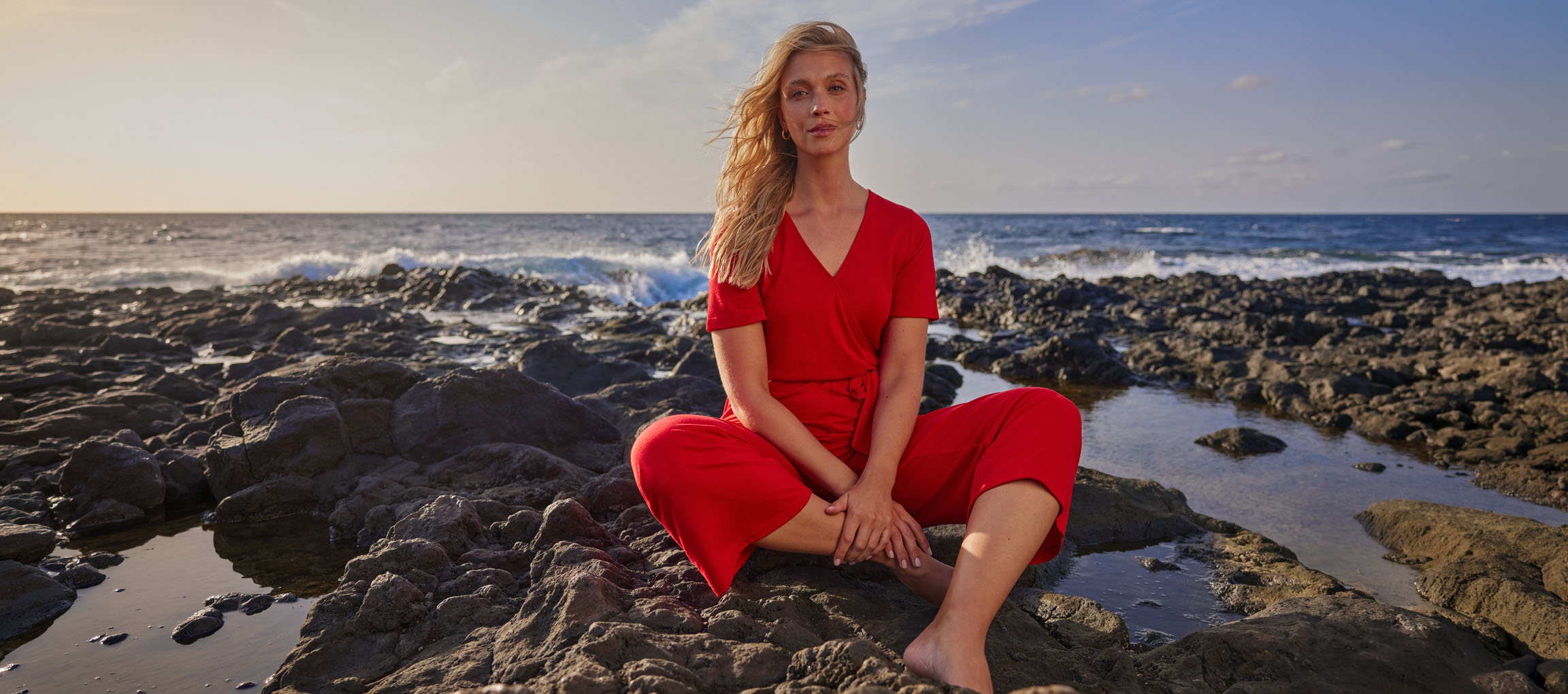 Femme en combinaison rouge assise sur une plage rocheuse au bord de la mer.