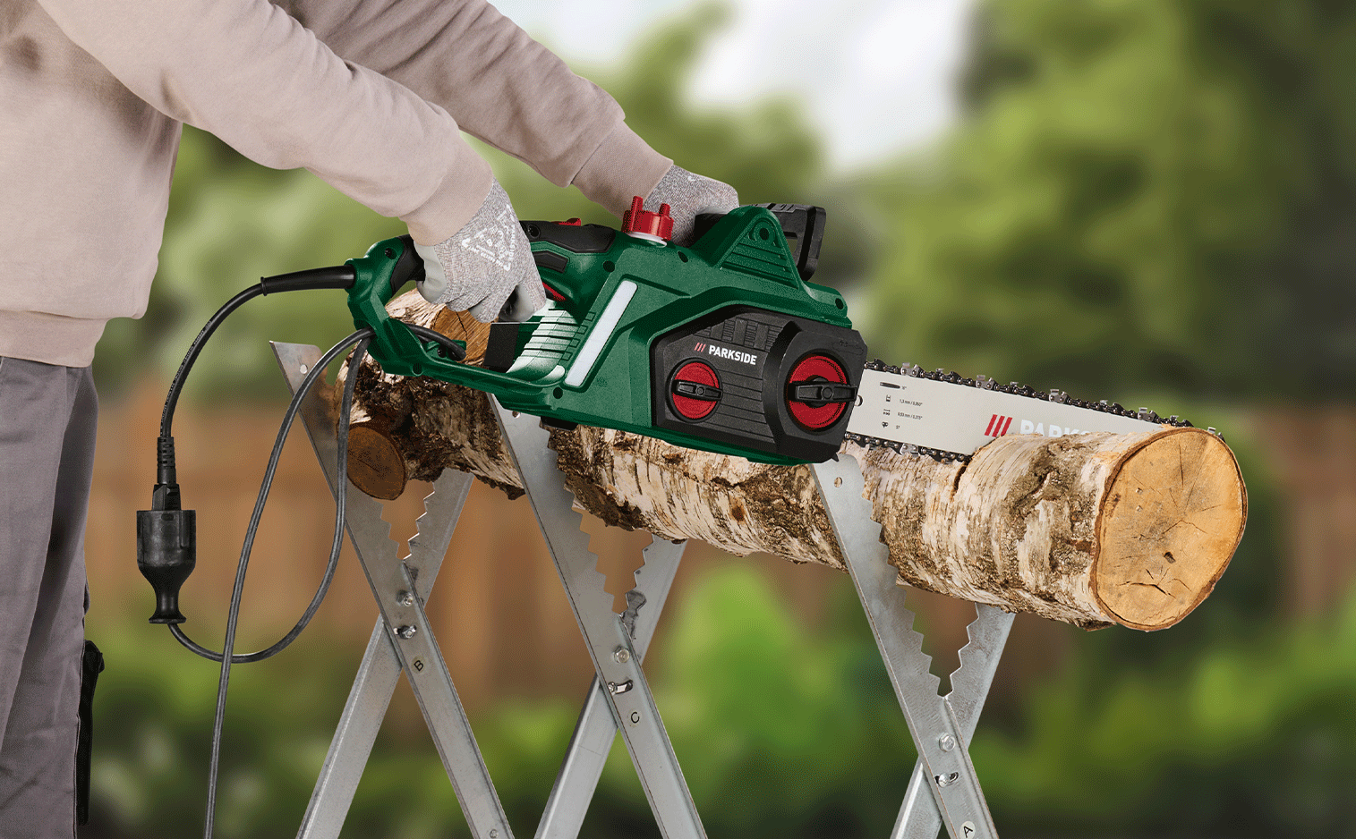 Tronçonneuse électrique coupant une bûche de bois sur un tréteau, tenue par des mains gantées.