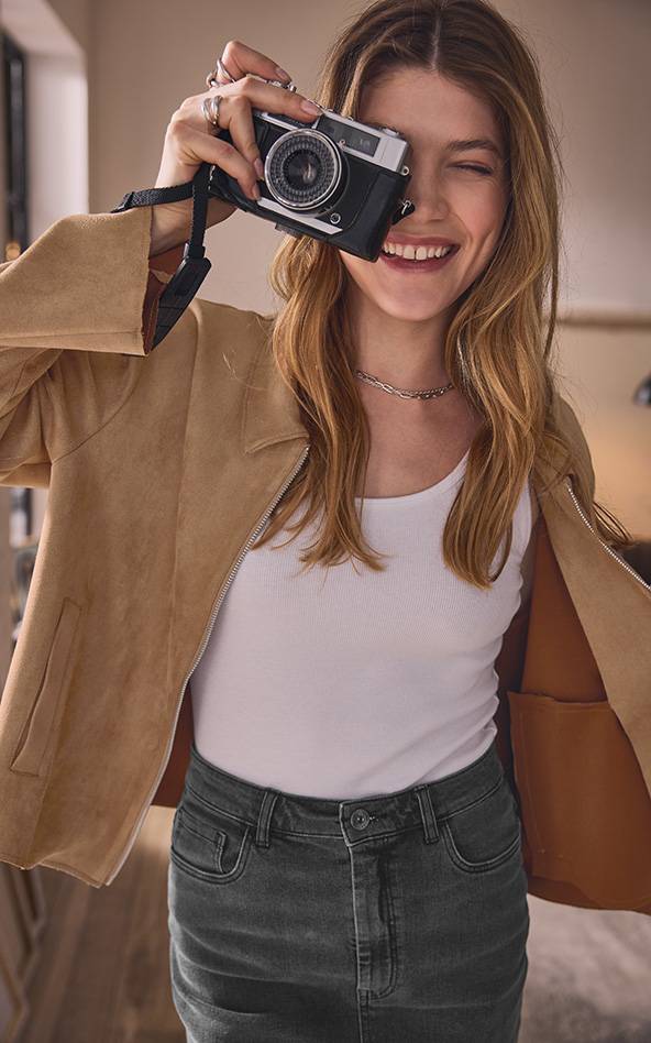 Une femme souriante tient un appareil photo vintage devant son œil, portant une veste en daim et un jean.