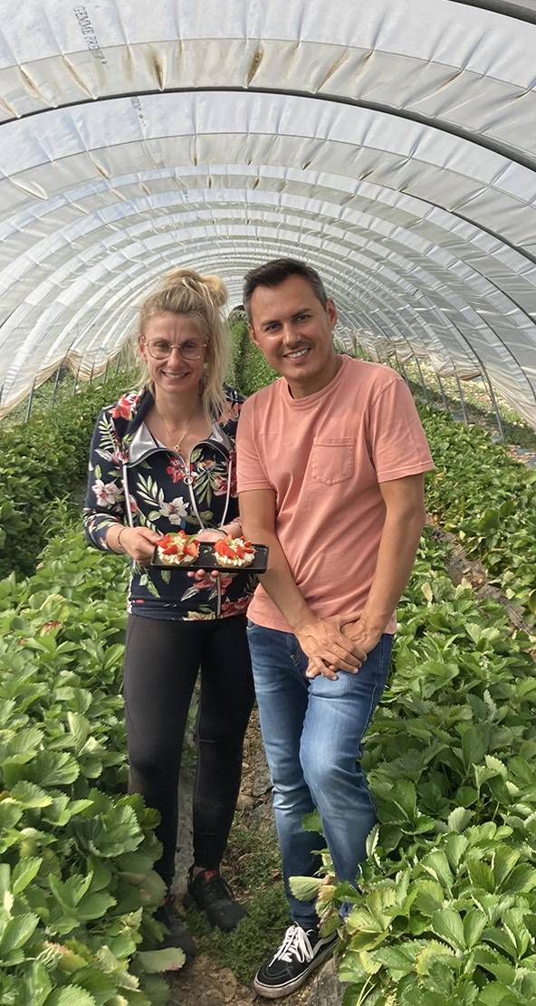 Deux personnes souriantes dans une serre à fraises, tenant des pâtisseries aux fraises.