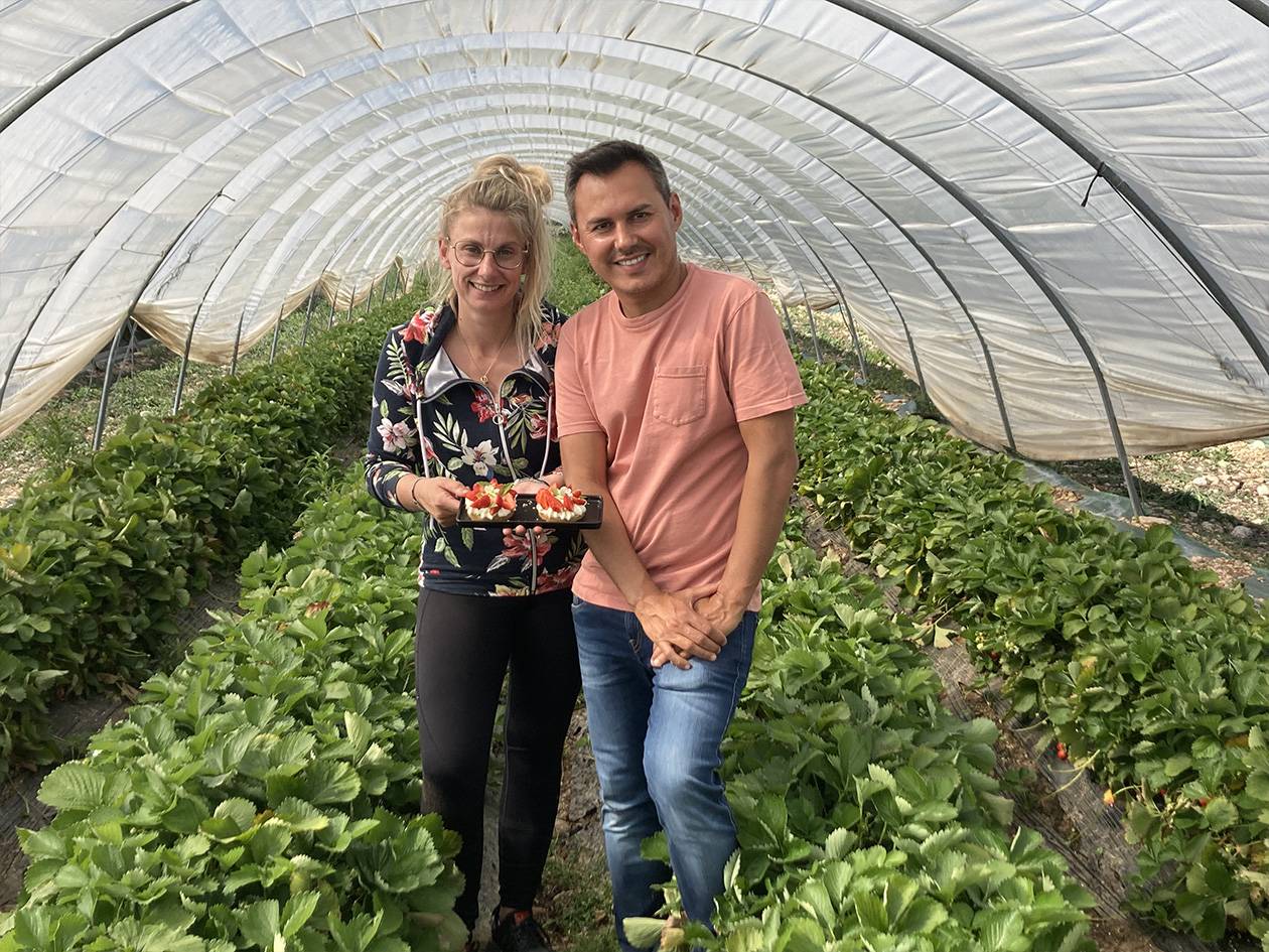 Deux personnes souriantes dans une serre à fraises, tenant une assiette de fraises.