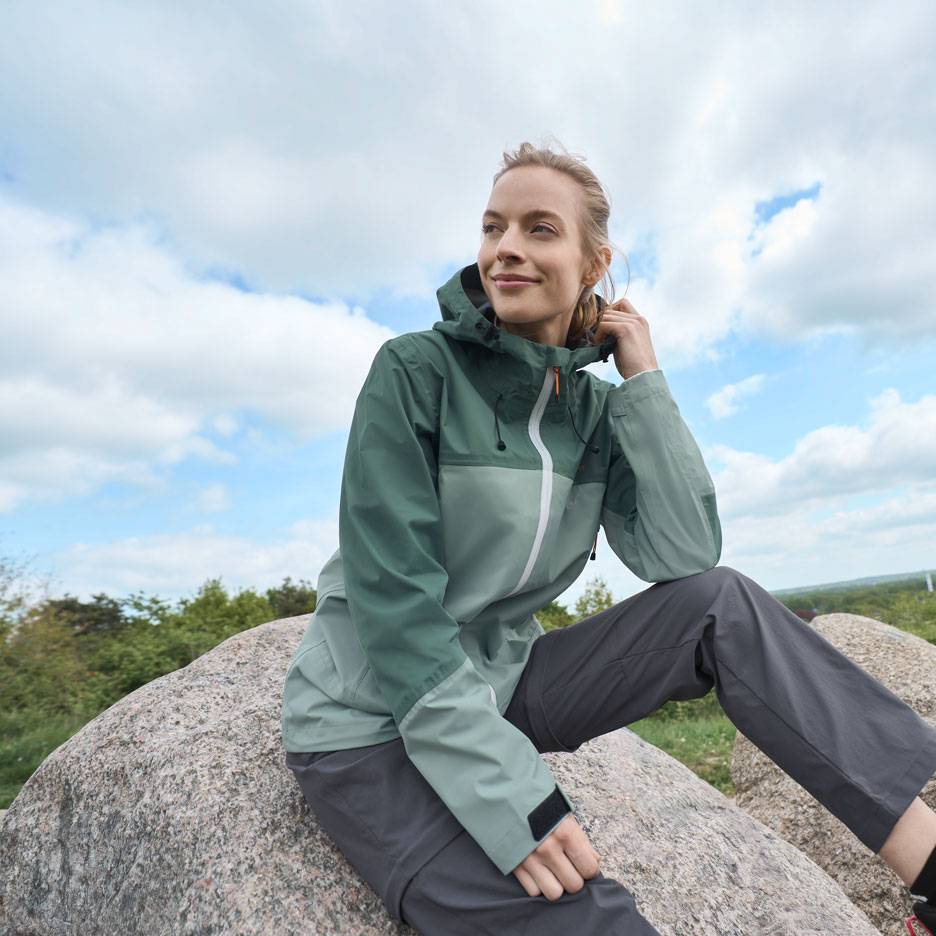 Femme en veste outdoor verte et pantalon gris assise sur un rocher, regardant au loin.