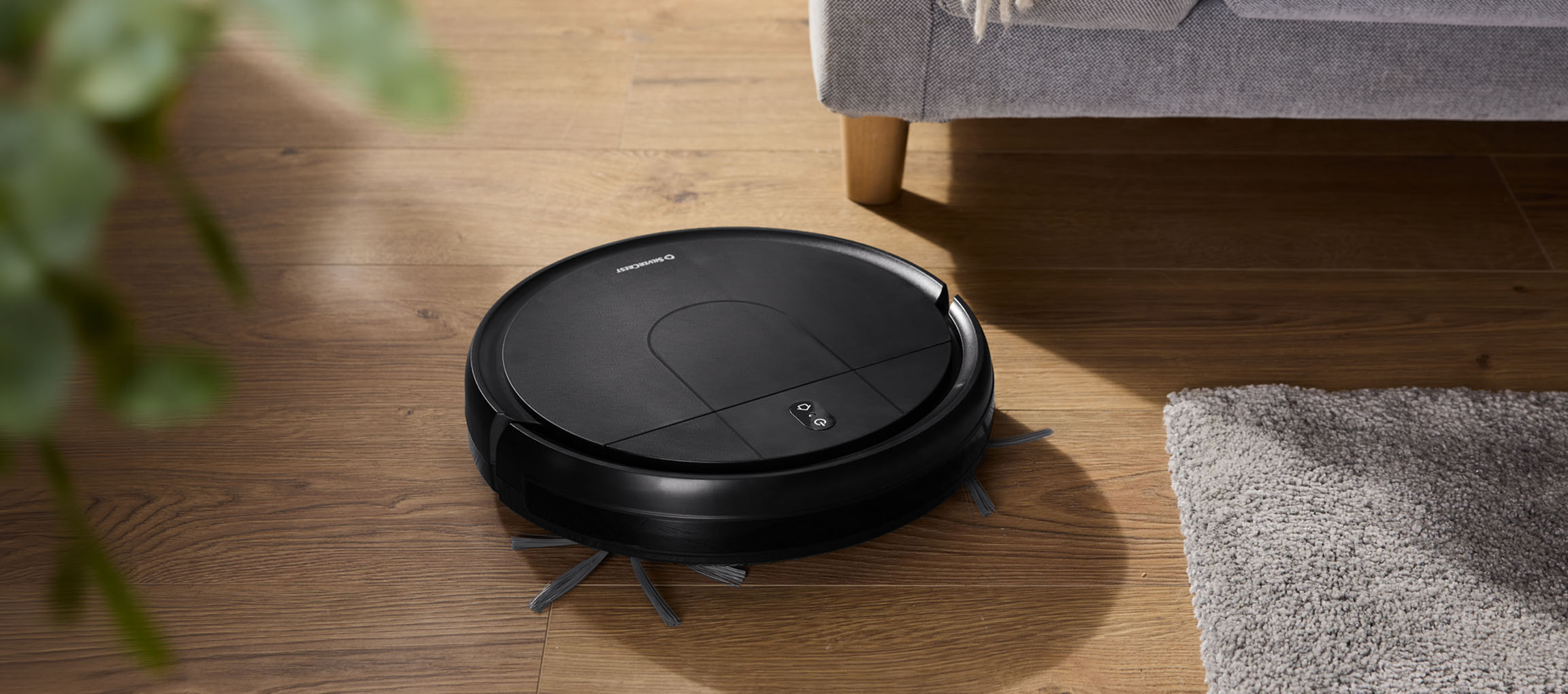 Aspirateur robot noir sur parquet à côté d'un canapé et d'un tapis.