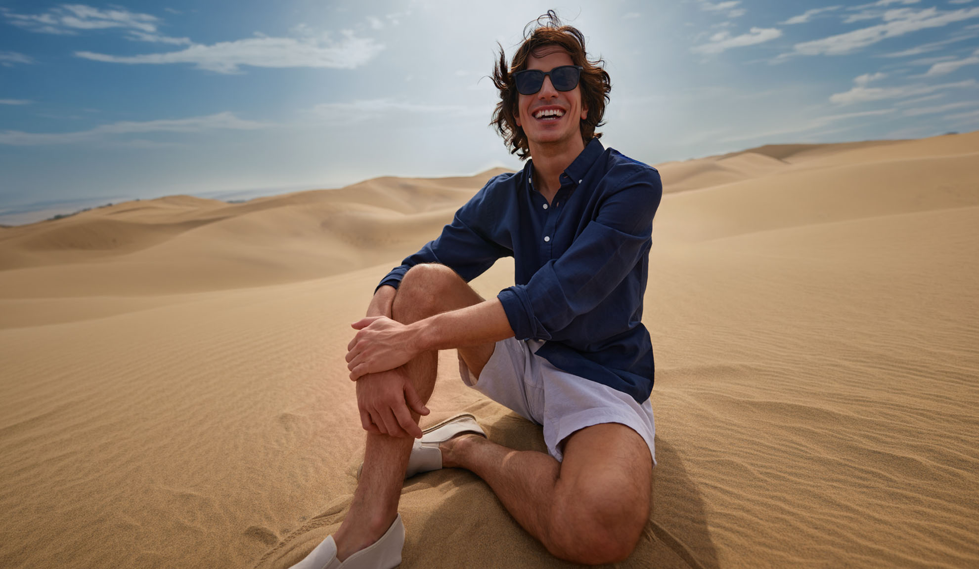 Homme en chemise bleu foncé et short clair assis sur des dunes de sable, souriant.