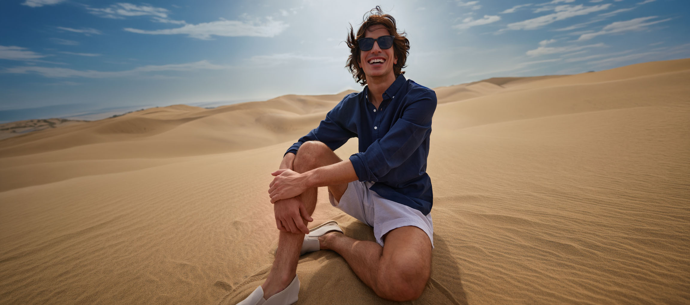 Homme en chemise bleu foncé et short assis dans les dunes de sable, souriant.