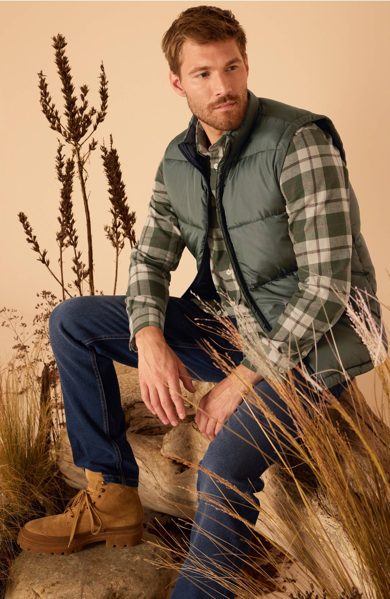Un homme porte un gilet matelassé vert, une chemise à carreaux et un jean, assis sur une bûche.