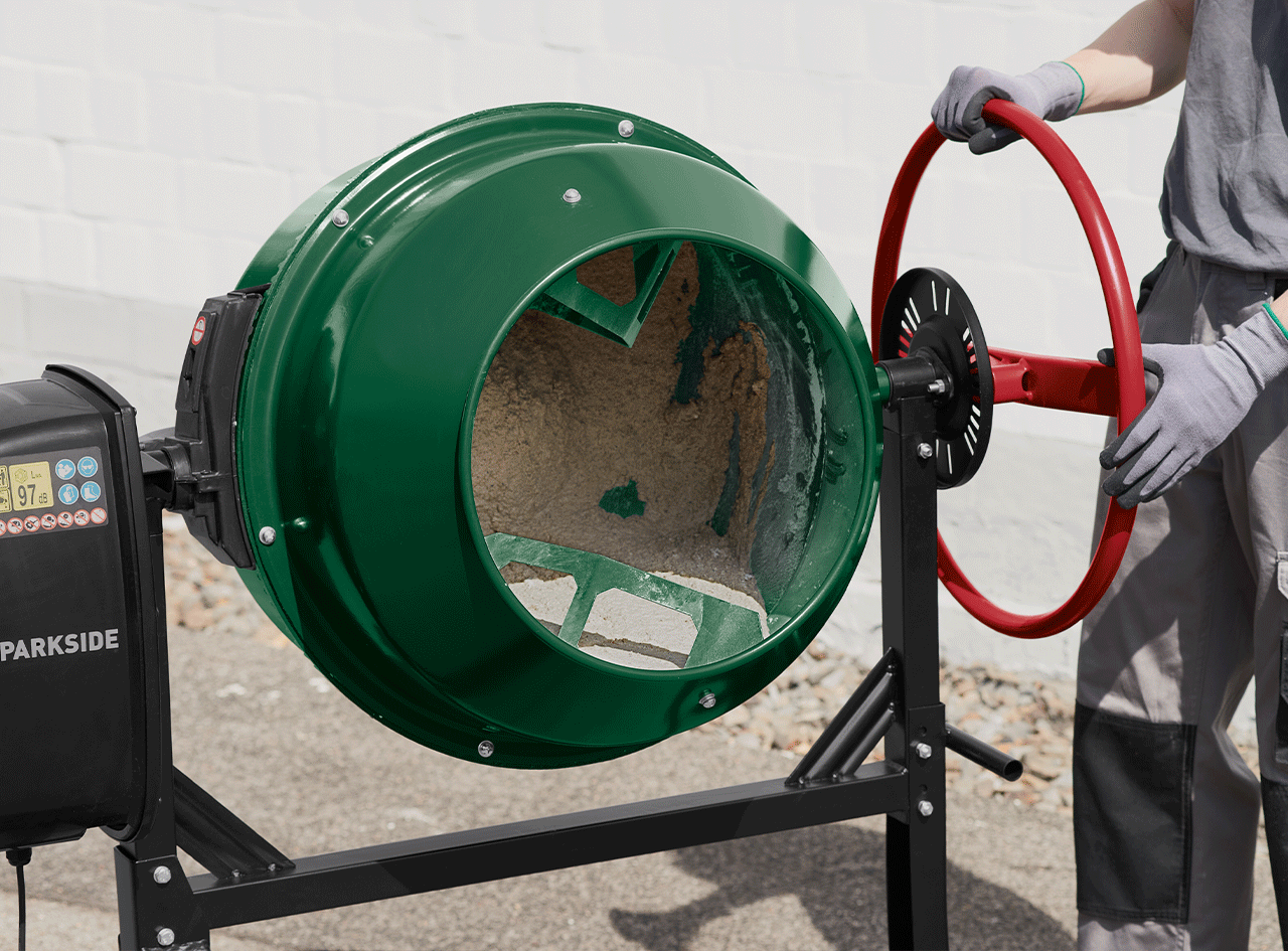 Bétonnière verte avec un ouvrier ajustant la roue rouge, sur un sol en béton.