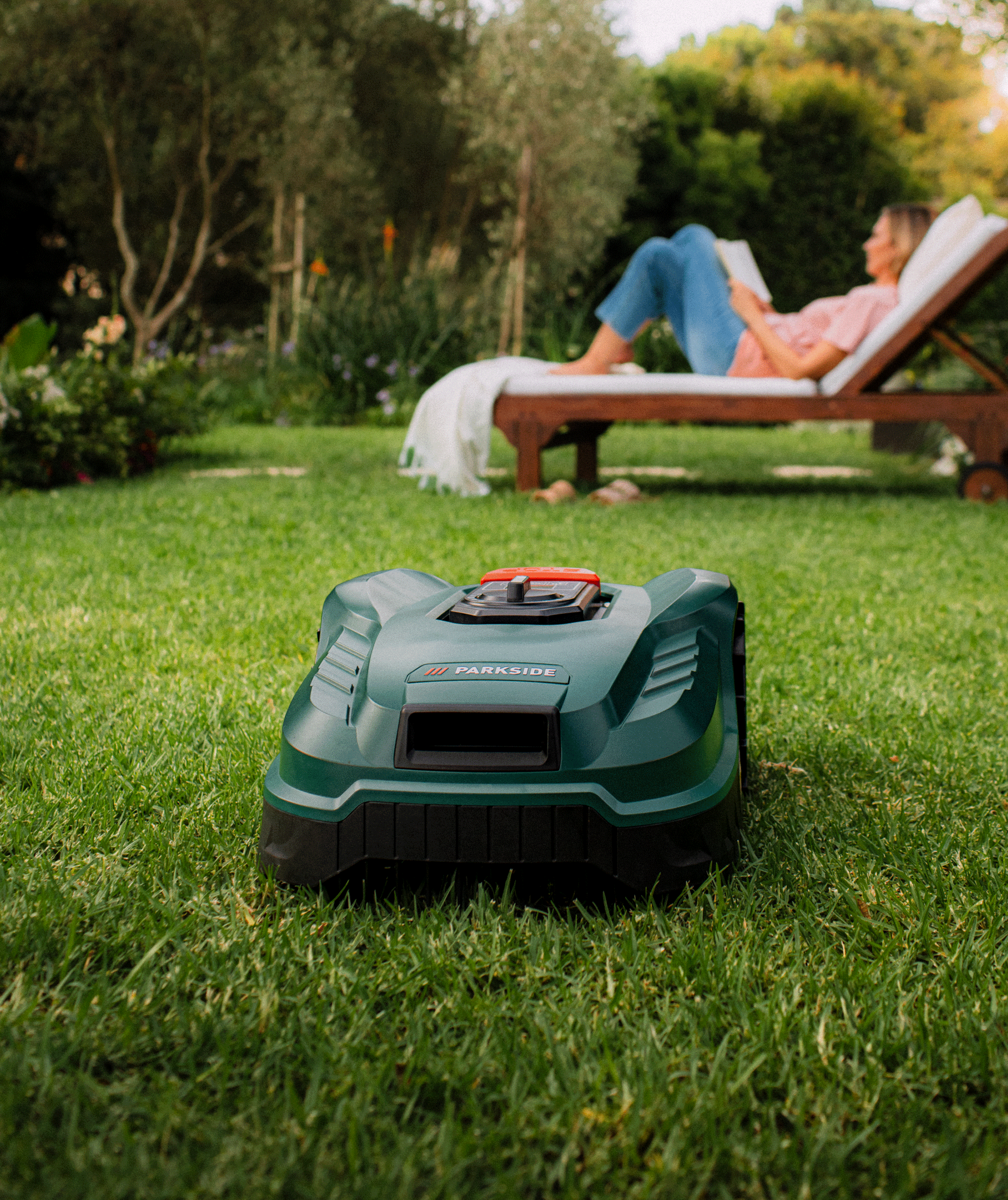 Un robot tondeuse sur une pelouse verte, avec une personne lisant sur une chaise longue en arrière-plan.
