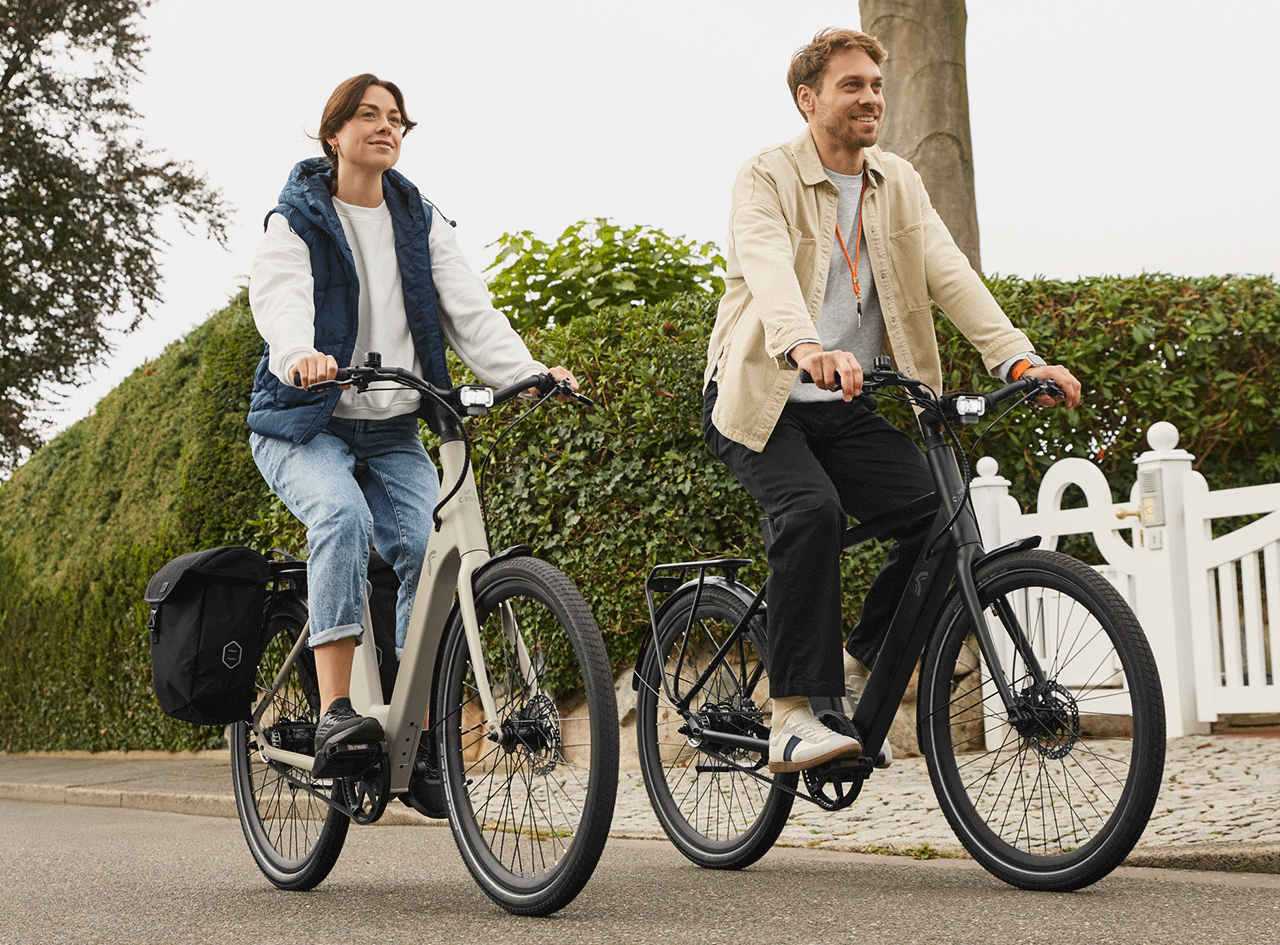 Deux personnes souriantes font du vélo électrique sur une route bordée de haies.