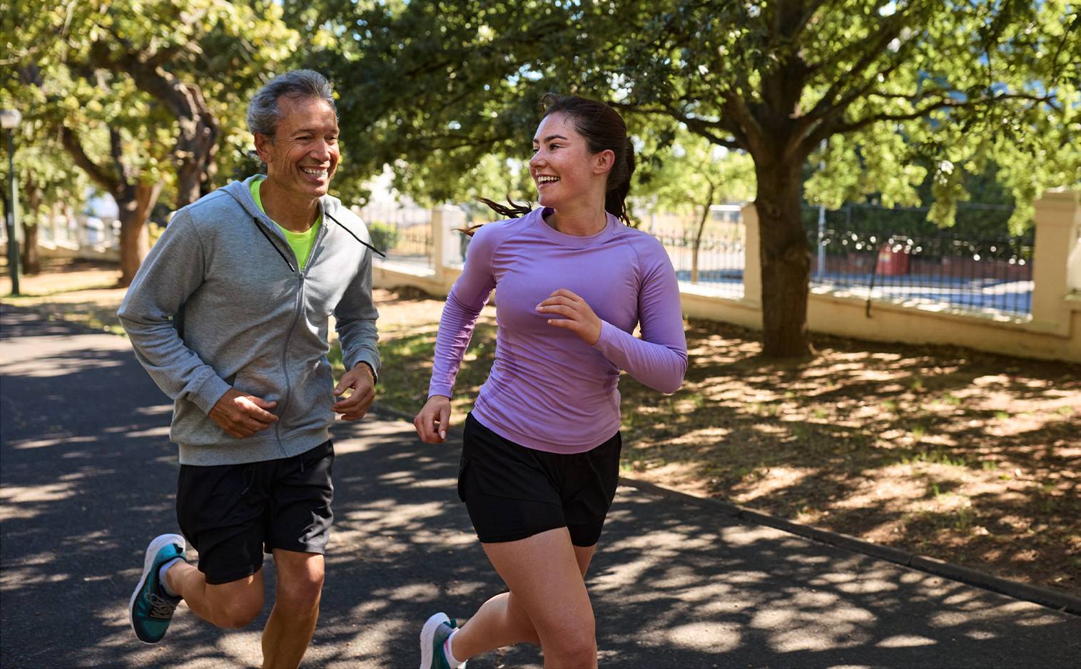 Un homme et une femme souriants en tenue de sport courent sur un chemin ombragé.