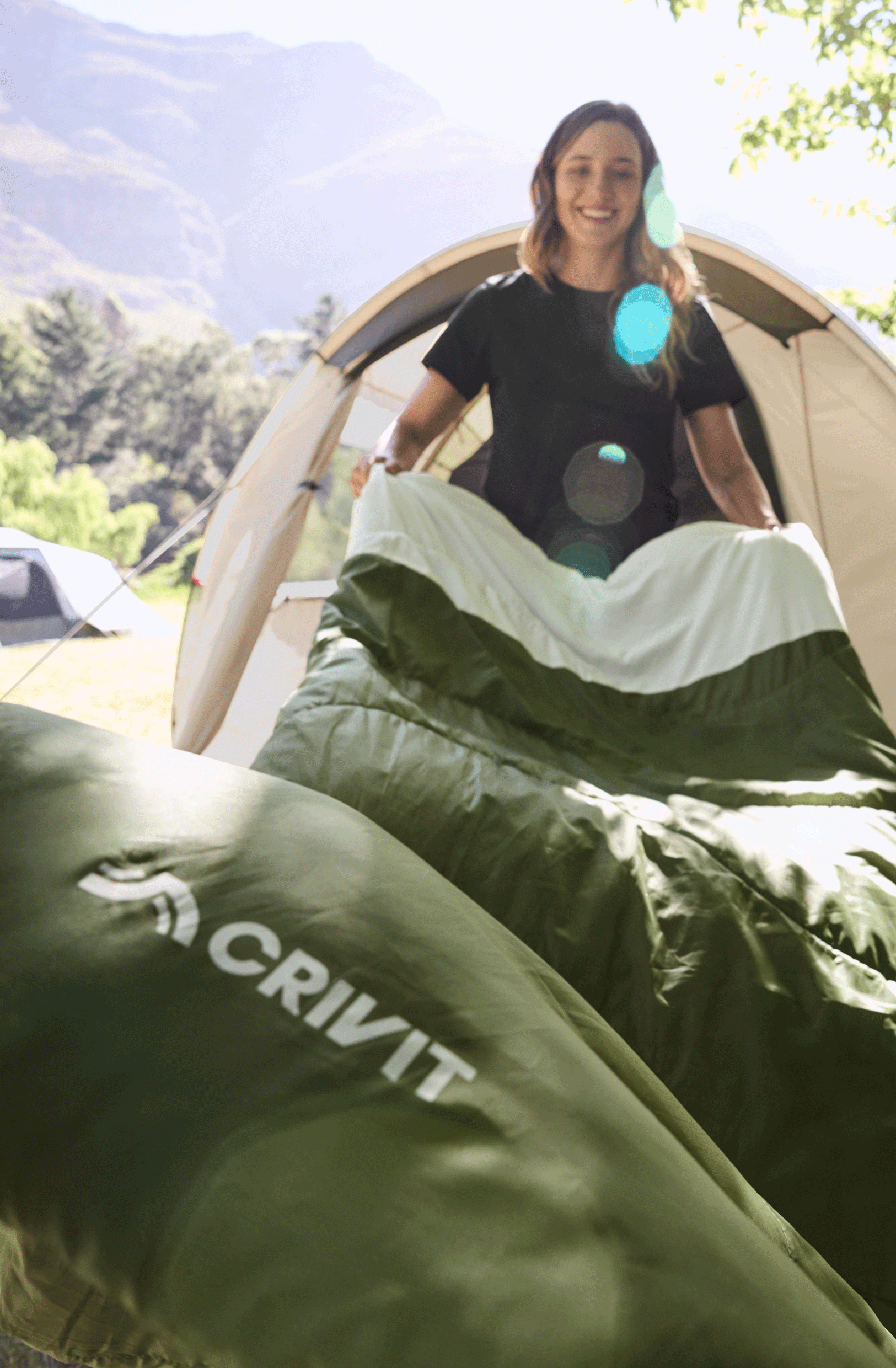 Une femme souriante dans une tente de camping, tenant un sac de couchage vert et blanc.