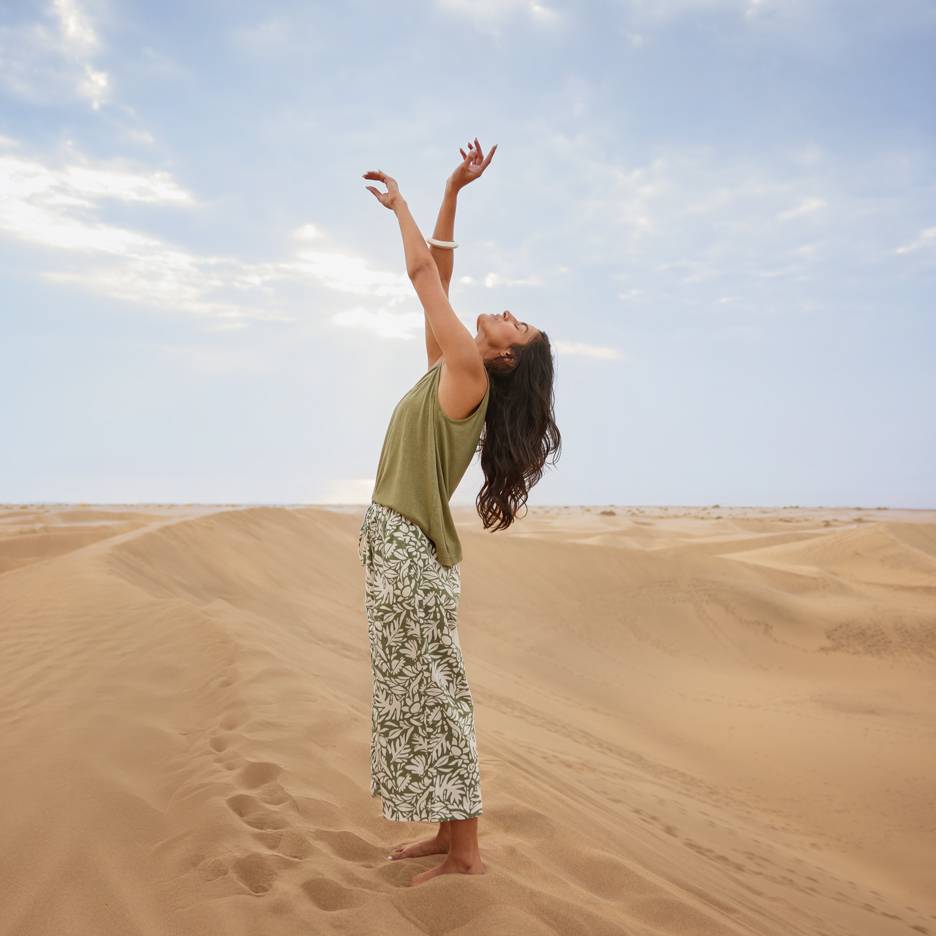Femme dans les dunes de sable avec un haut vert et un pantalon à motifs, bras levés.