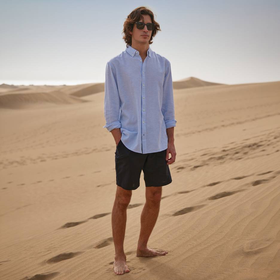 Homme en chemise bleue et short noir, pieds nus sur des dunes de sable.