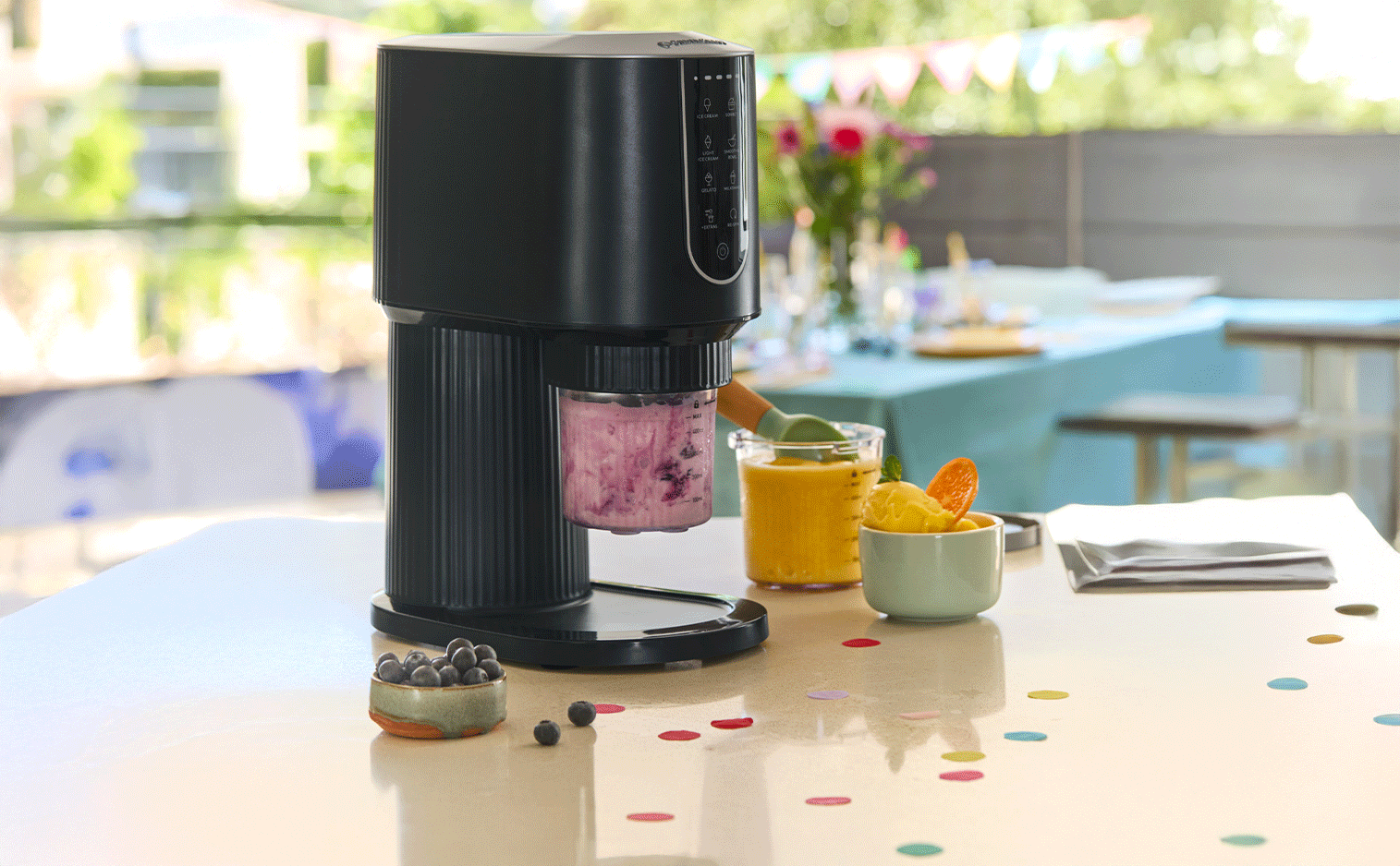Machine à glace noire avec un pot de glace aux baies, un verre de smoothie et un bol de sorbet sur une table de fête.