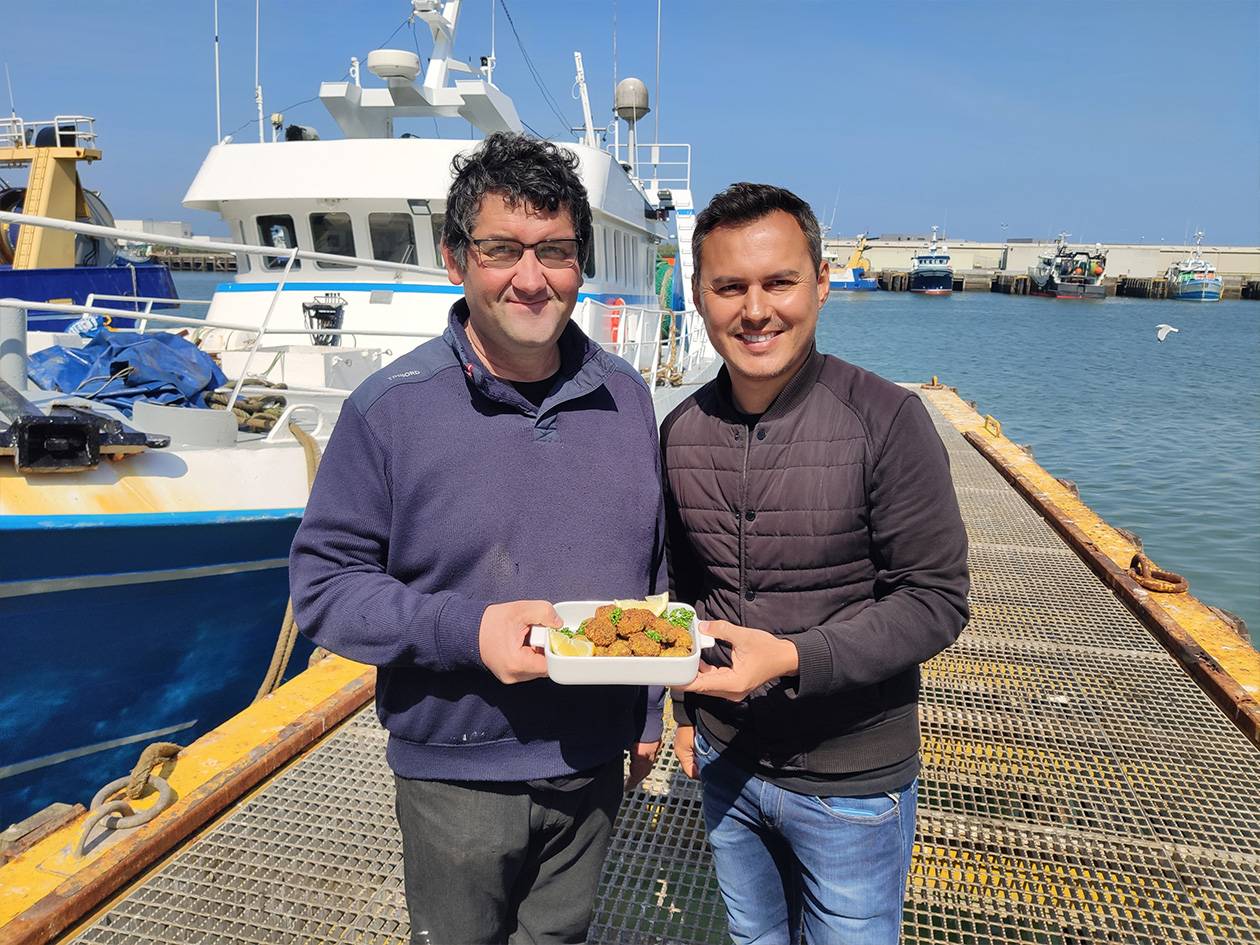 Deux hommes souriants tenant un plat de poisson frit sur un quai de pêche avec des bateaux.