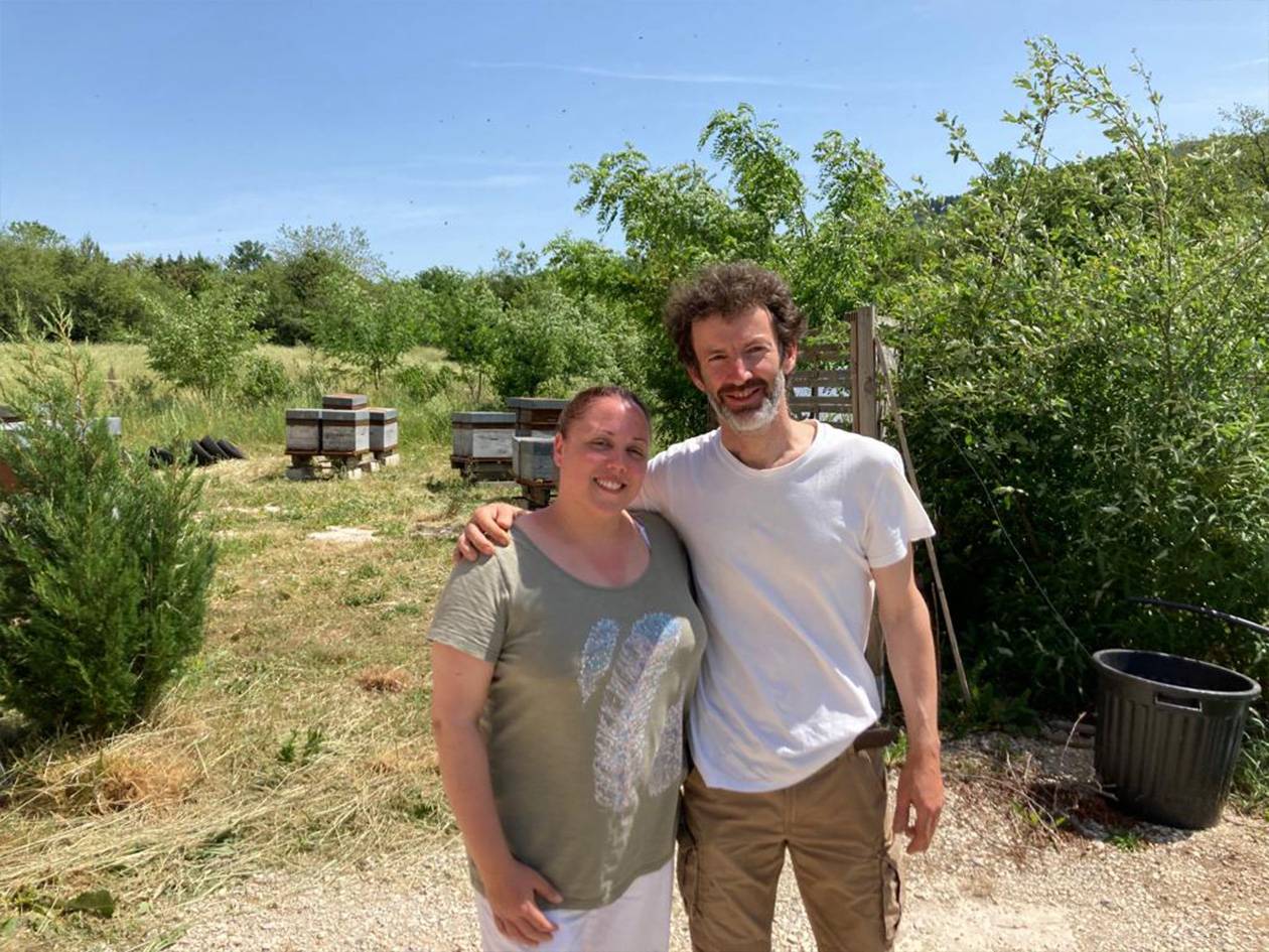 Un homme et une femme souriants posent devant des ruches dans un champ ensoleillé.