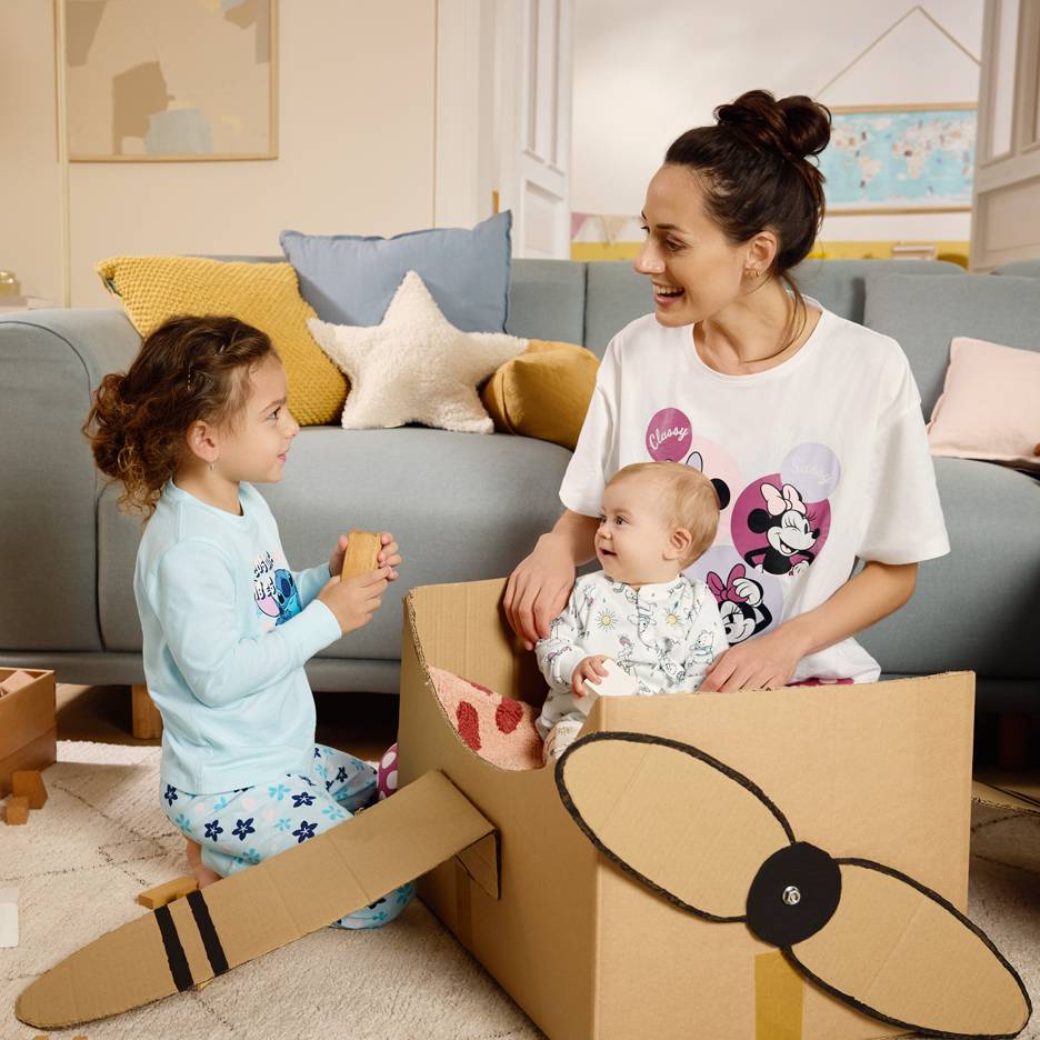 Maman et enfants jouant avec une boîte en carton, la maman portant un t-shirt à personnages de dessins animés.