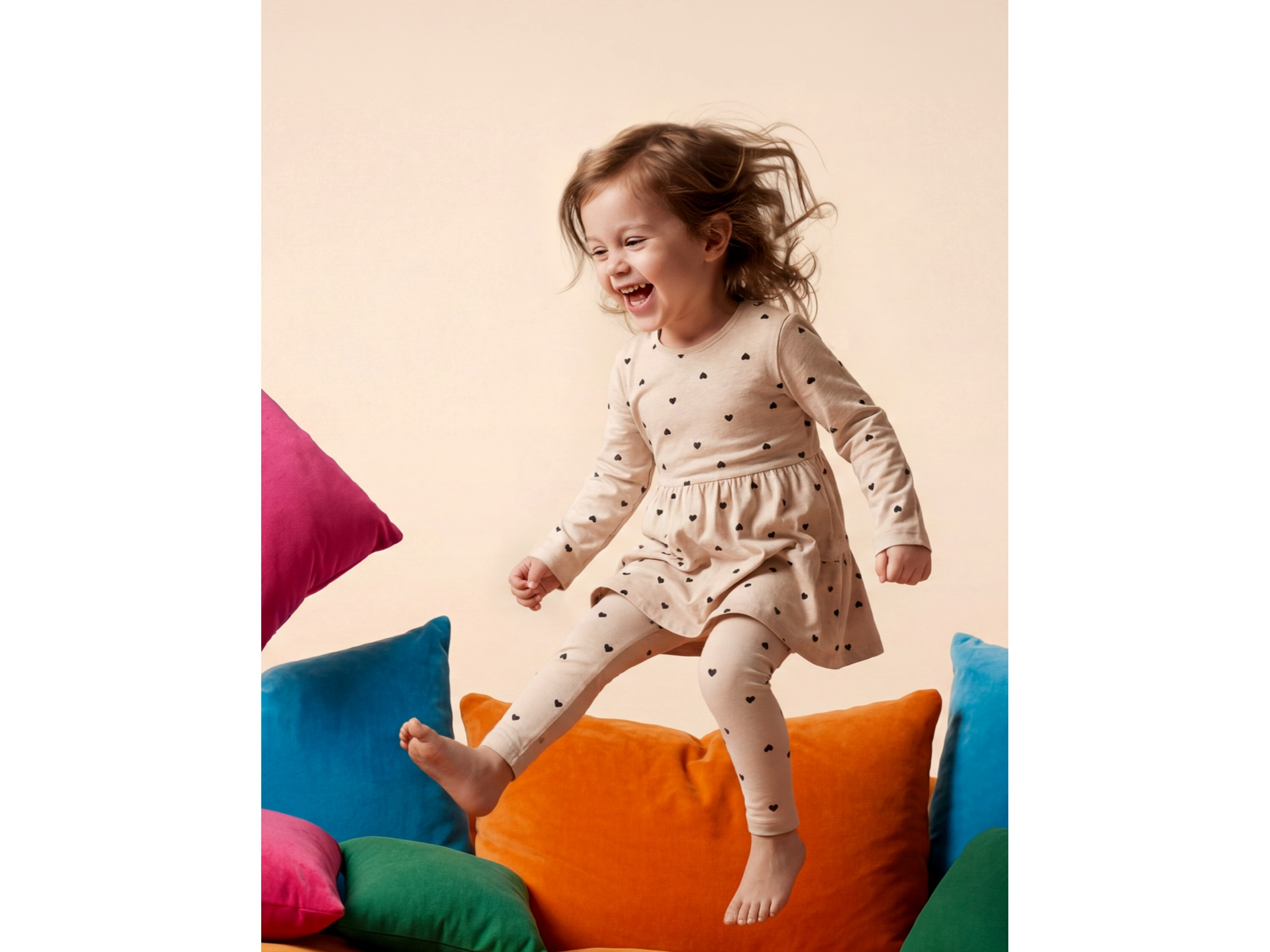Petite fille joyeuse en robe et leggings à cœurs LUPILU, sautant sur des coussins colorés.
