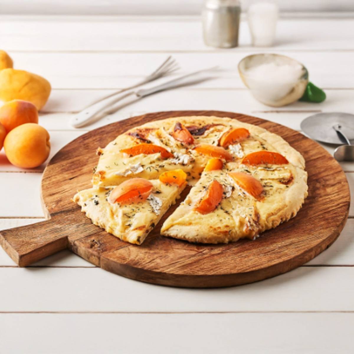 Une pizza à la crème, aux abricots et au brie, coupée en parts, servie sur une planche en bois.