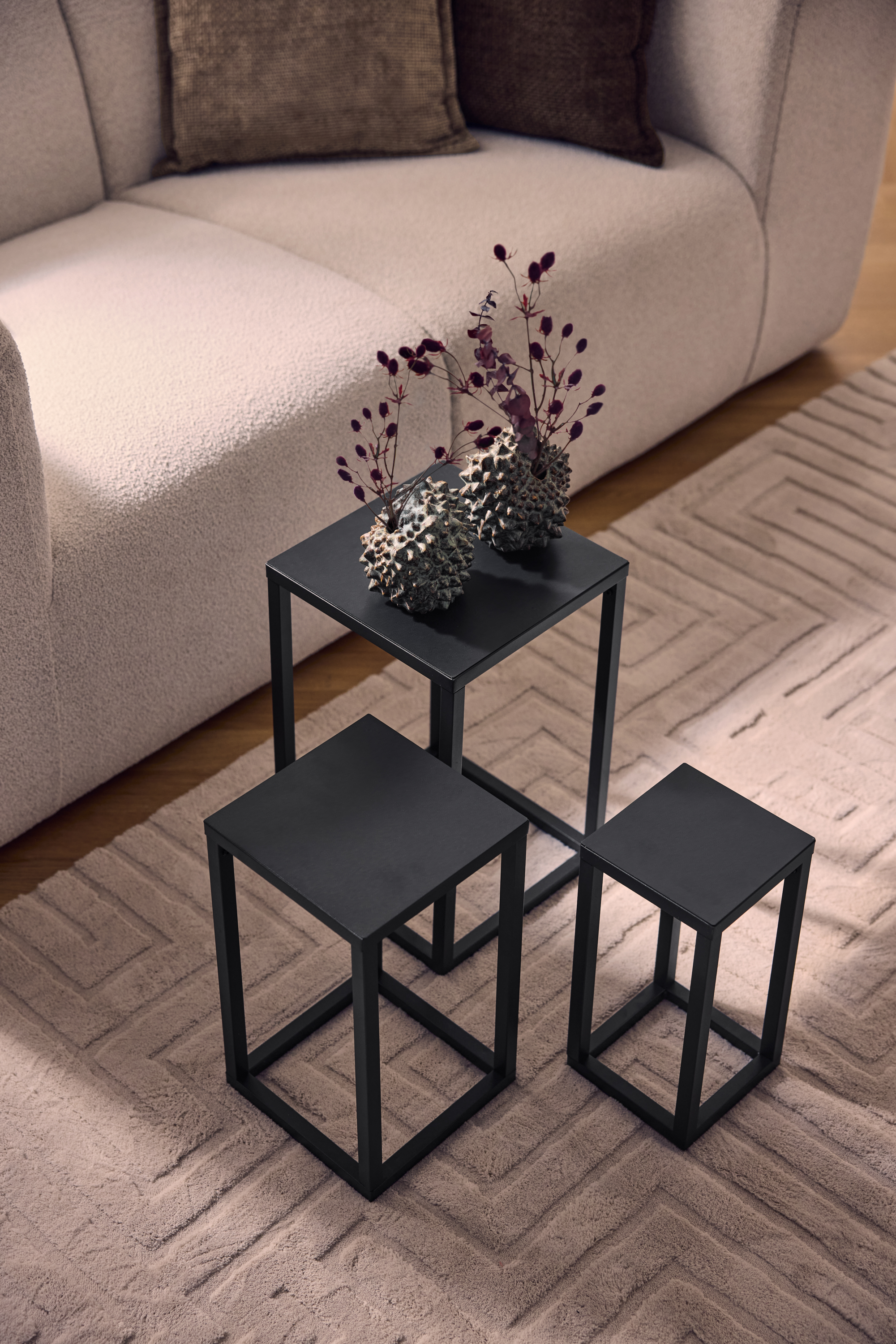 Trois tables basses en métal noir sur un tapis beige à motif géométrique, à côté d'un canapé clair.