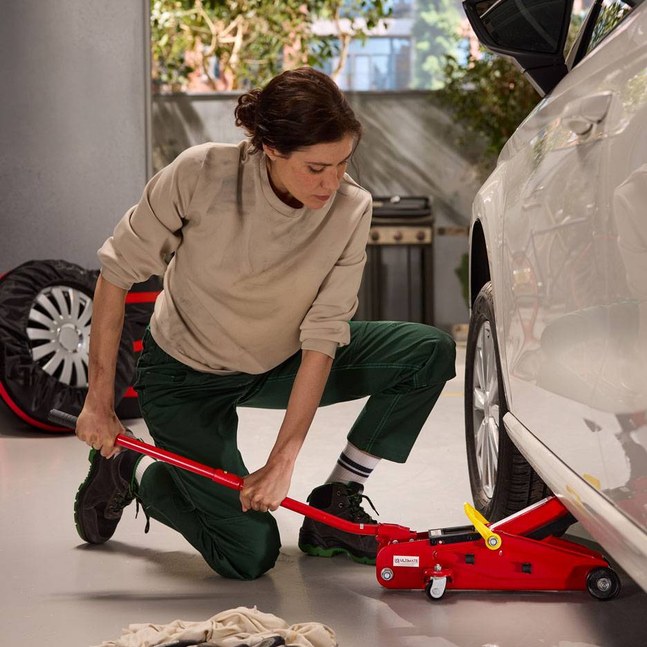 Femme utilisant un cric hydraulique rouge pour soulever une voiture blanche, avec une roue de secours à proximité.