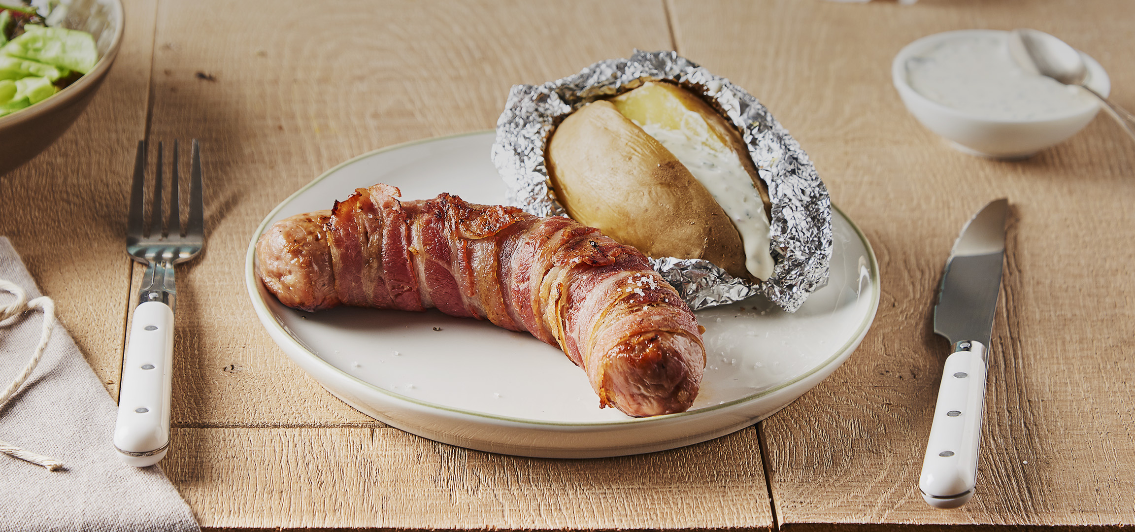 Saucisse enroulée de bacon avec une pomme de terre au four et de la salade.