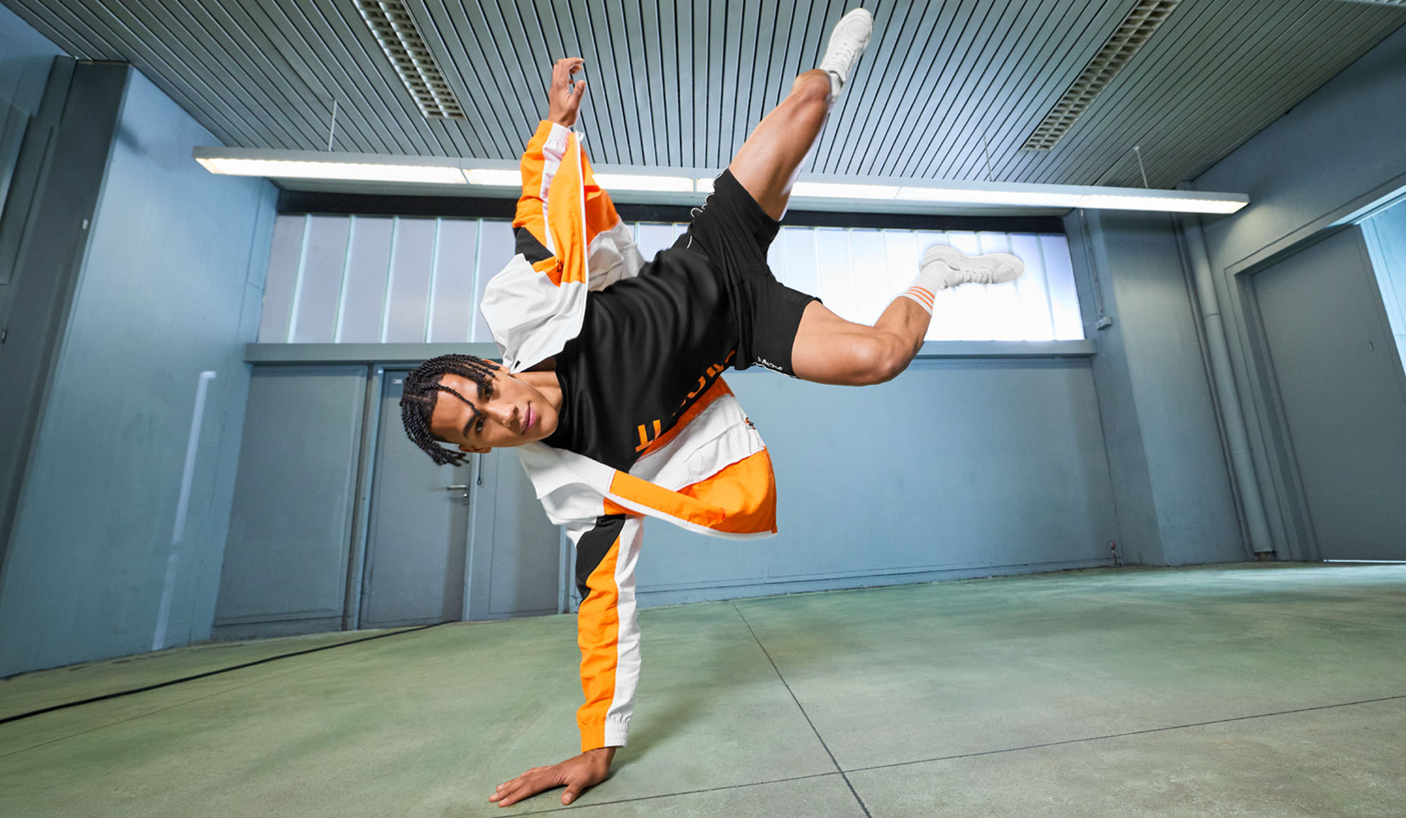Homme en tenue de sport, t-shirt noir, short et veste orange et blanche, exécutant un mouvement de danse.