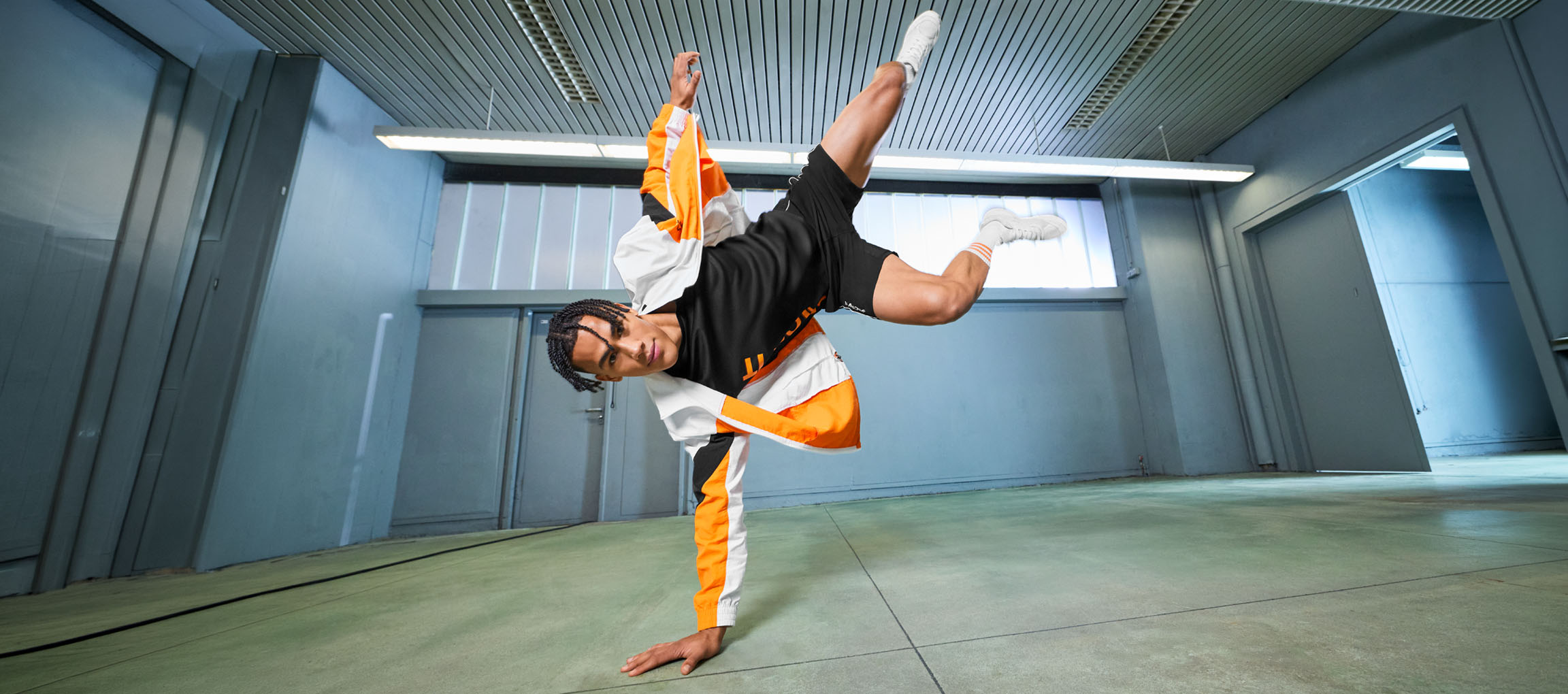Homme en tenue de sport, t-shirt noir et veste orange et blanche, dansant dans une pièce.