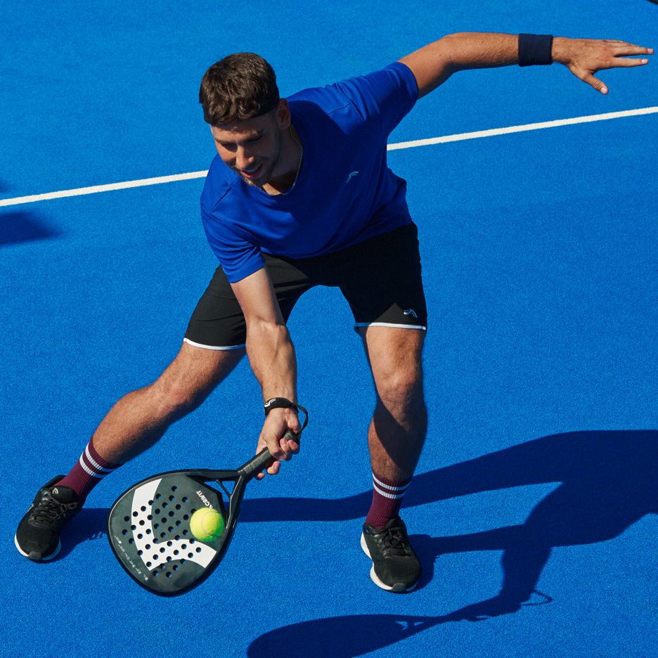 Homme en t-shirt bleu et short noir jouant au padel avec raquette et balle.