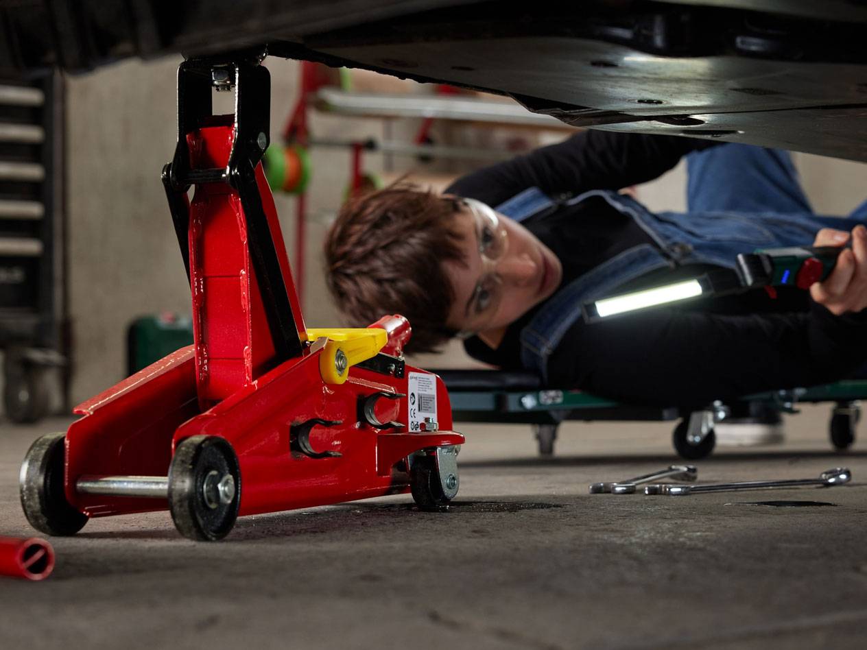 Cric hydraulique rouge et femme sous une voiture avec lampe de travail et outils.