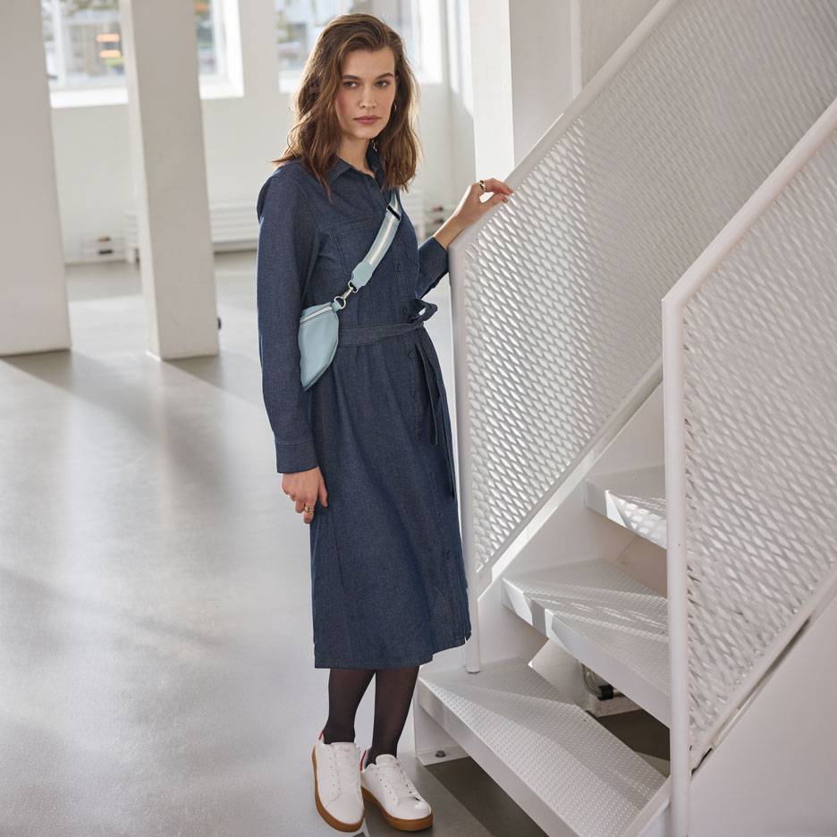 Femme en robe chemise en jean, collants noirs et baskets blanches, avec un sac banane bleu clair.