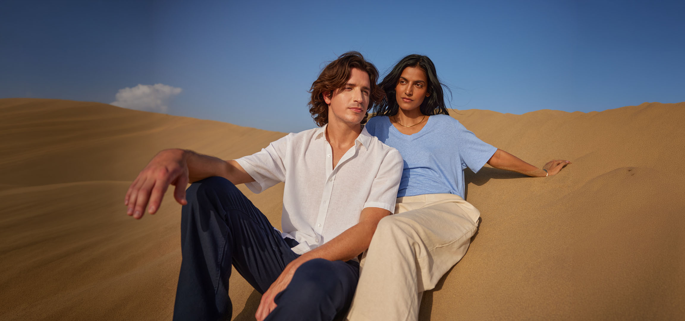 Homme et femme assis sur des dunes de sable, portant des chemises et pantalons en lin.