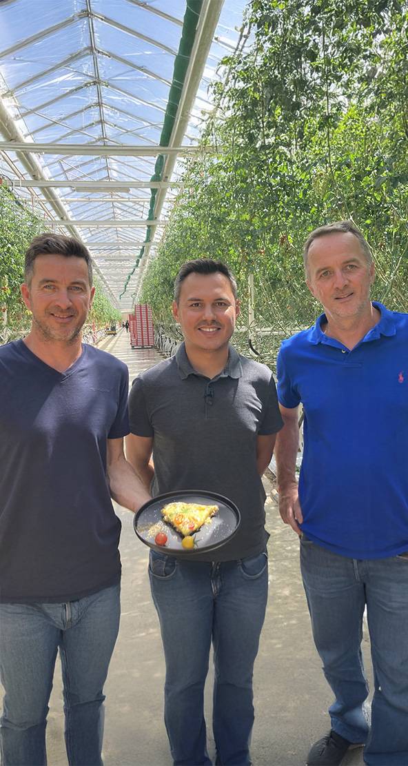 Trois hommes souriants dans une serre, l'un tenant une assiette de nourriture.