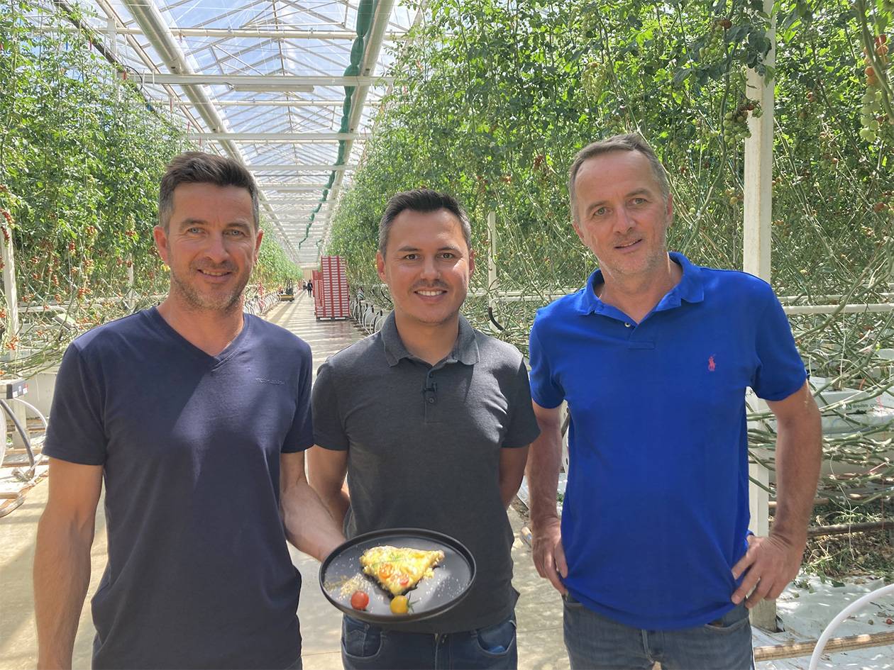 Trois hommes souriants dans une serre de tomates, l'un tenant une assiette de pizza.