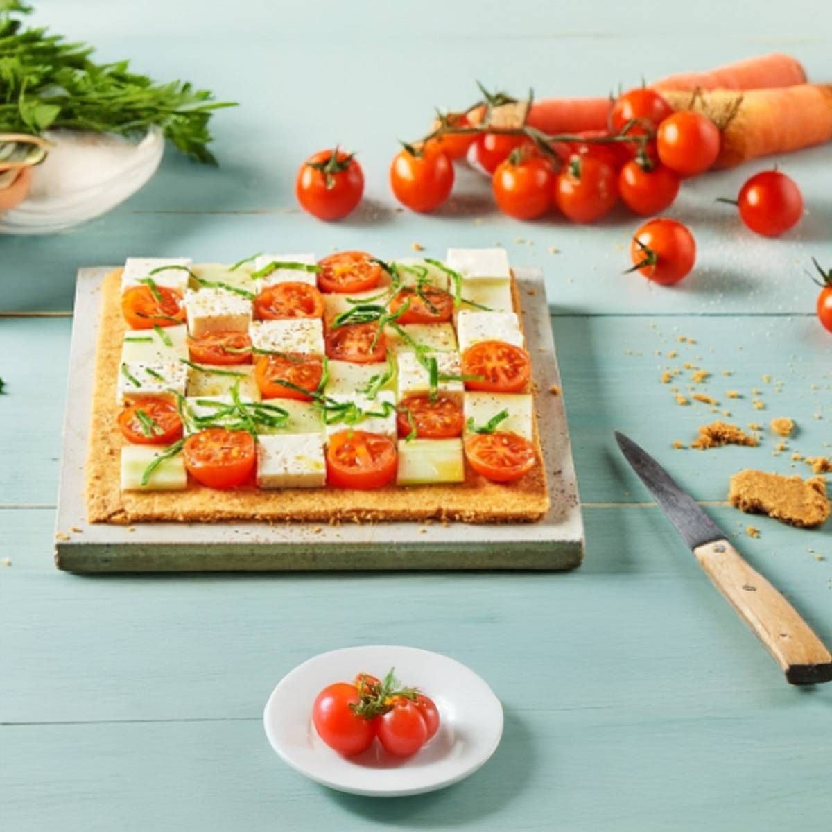 Une tarte salée avec des tomates cerises, du fromage et du concombre sur une table bleue.