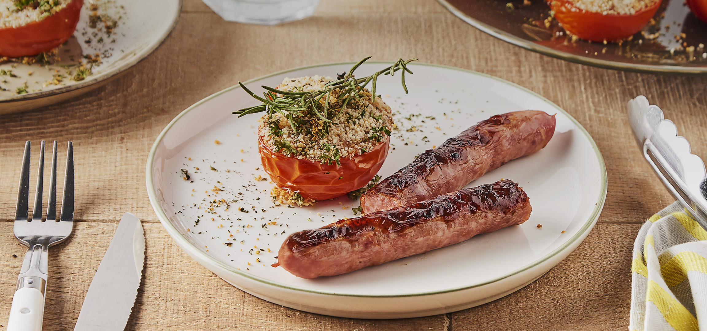 Saucisses grillées et tomate farcie aux herbes sur une assiette blanche.