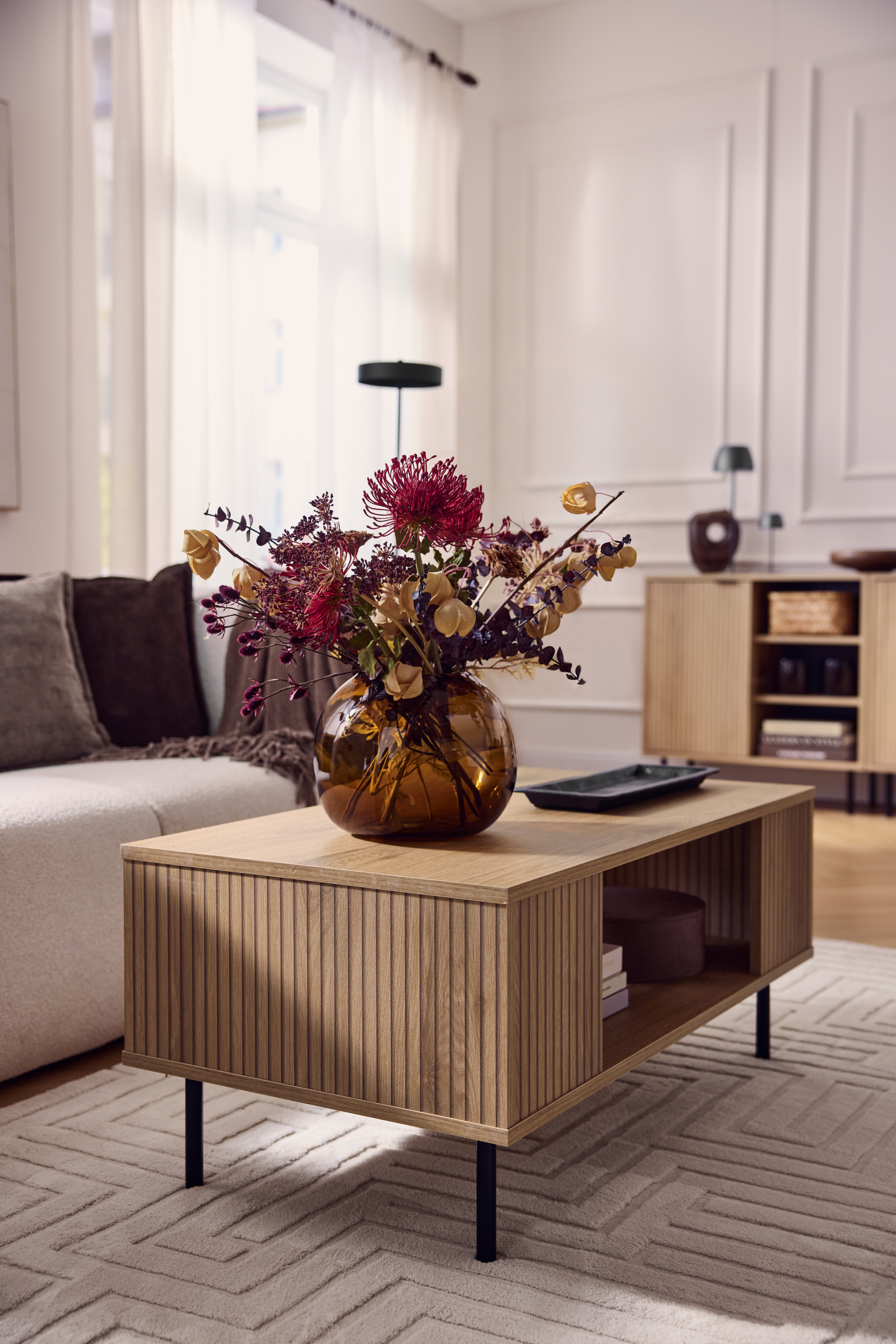 Table basse en bois avec façade cannelée, vase de fleurs séchées, canapé beige.