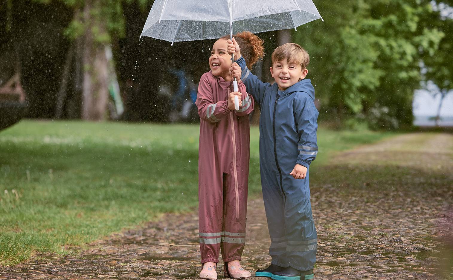 Deux enfants souriants en combinaisons de pluie sous un parapluie transparent sous la pluie.