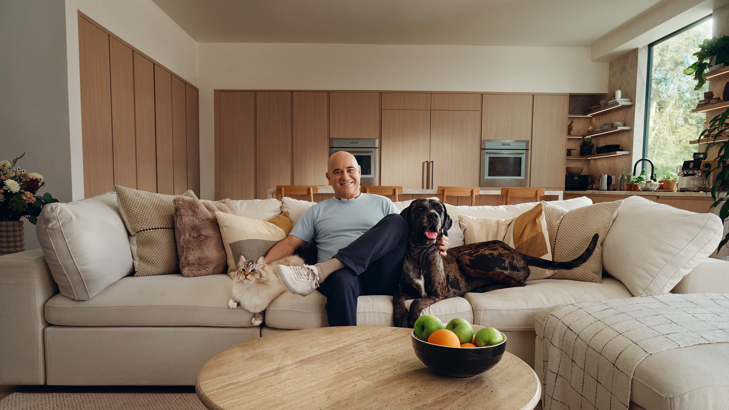 Homme sur canapé avec chat et chien, coupe de fruits sur table basse.